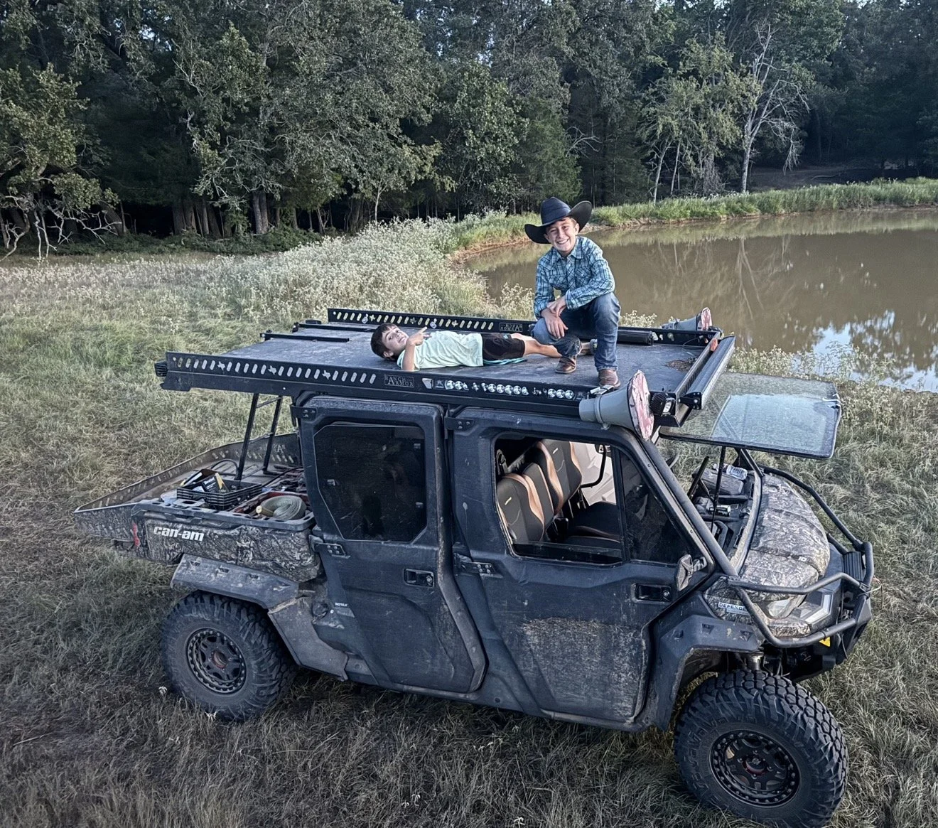 Two children, one lying down and the other kneeling, on top of an ATV near a pond in a wooded area.