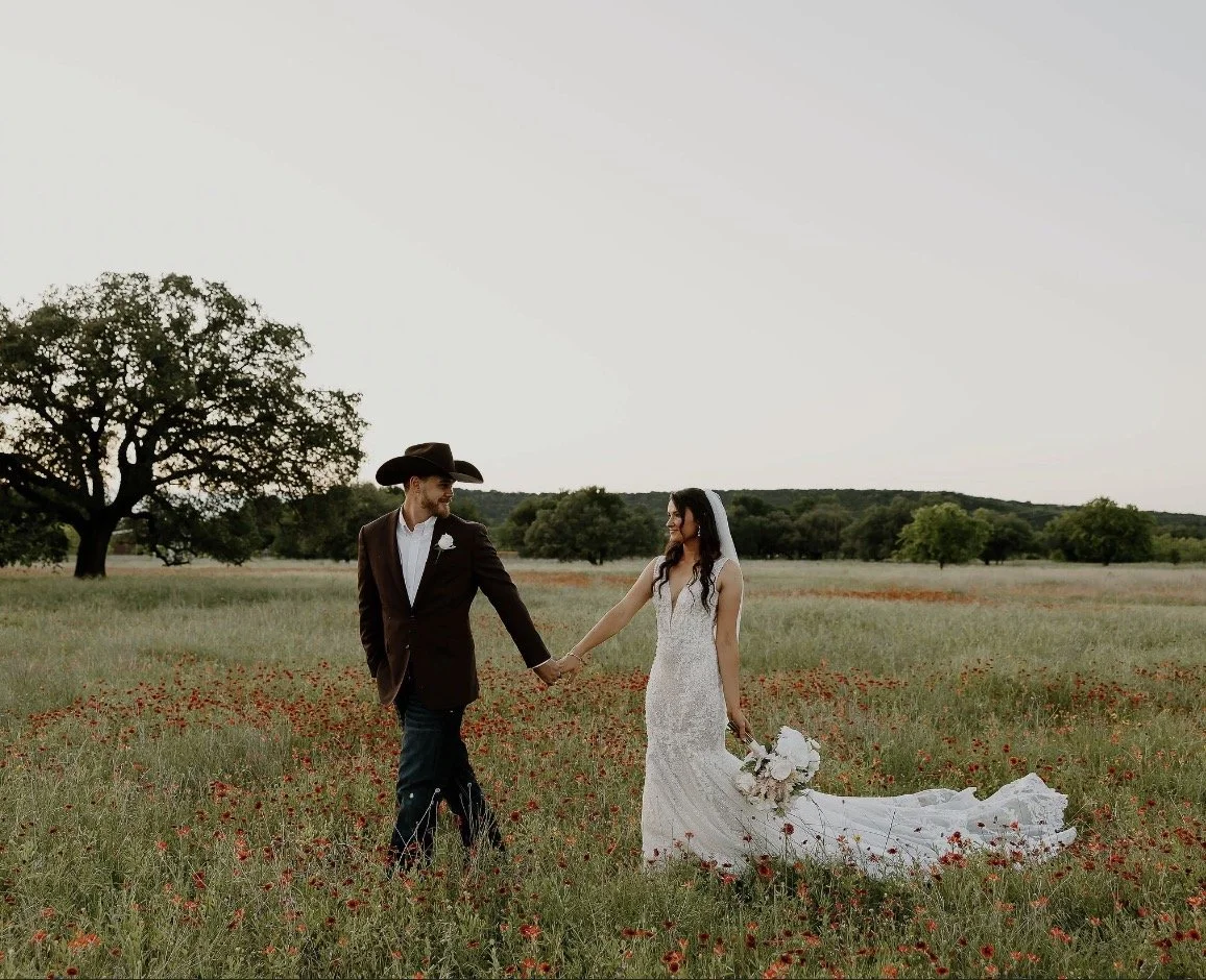 A bride and groom holding hands in a field with green grass and red flowers, with trees and a clear sky in the background.