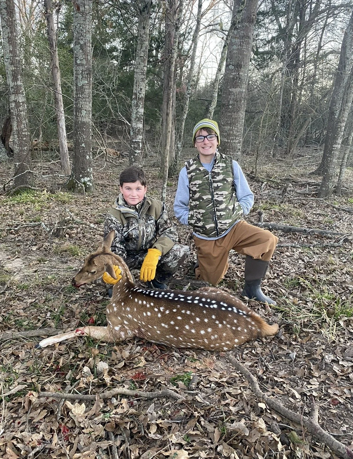 Two boys in camouflage clothing and rubber boots in a wooded area, one sitting and the other standing, with a small deer lying on the ground in front of them.