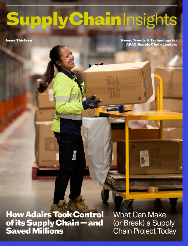 Cover image of Supply Chain Insights Digital Magazine. A woman in a lime green safety jacket and black gloves smiling while holding a cardboard box in a warehouse. There are other boxes and a yellow cart nearby.