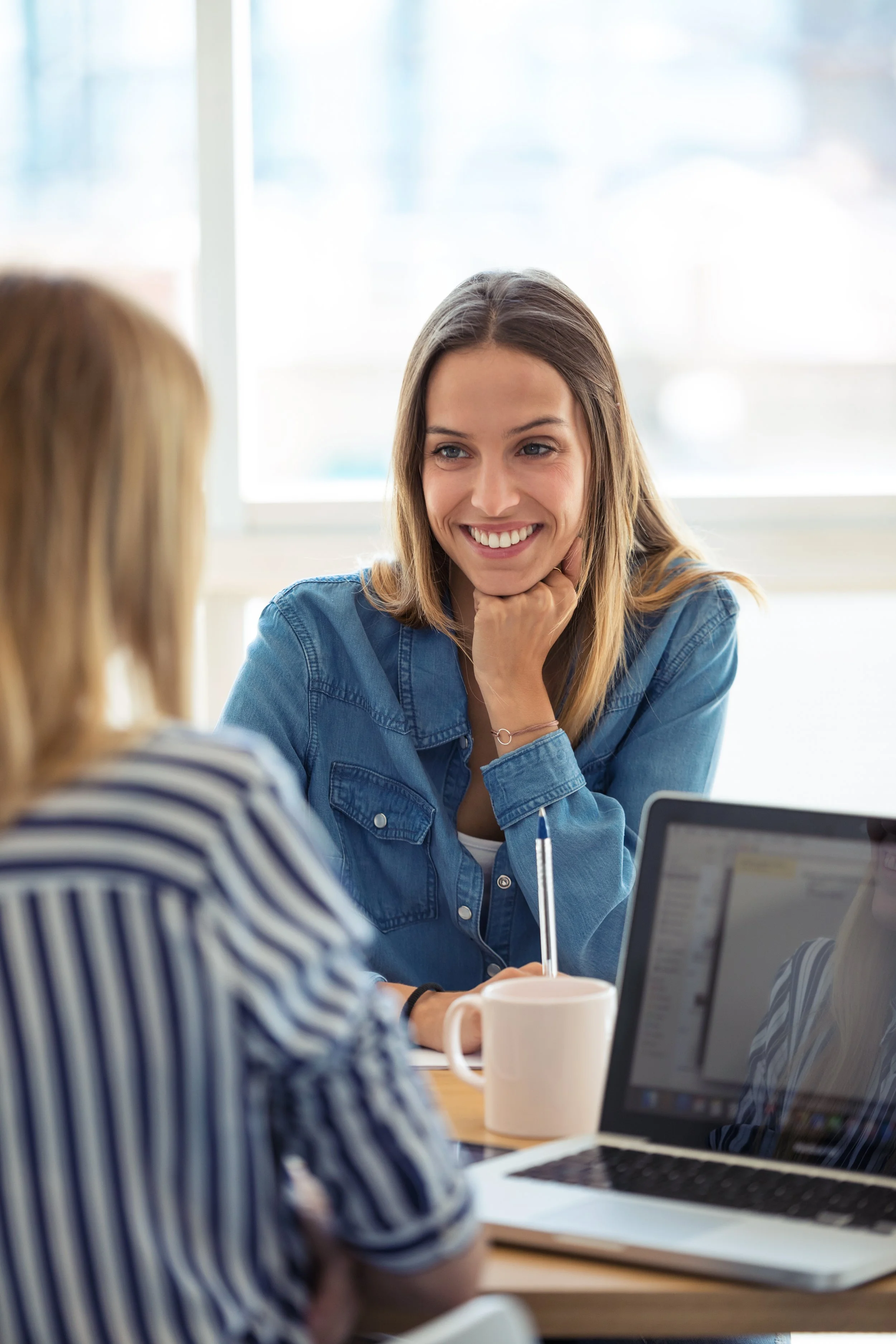To kvinder sidder ved et bord, og den ene af dem smiler. Den anden kvinde kan ikke ses tydeligt. Der er en kop, en åben computer og en pære på bordet.