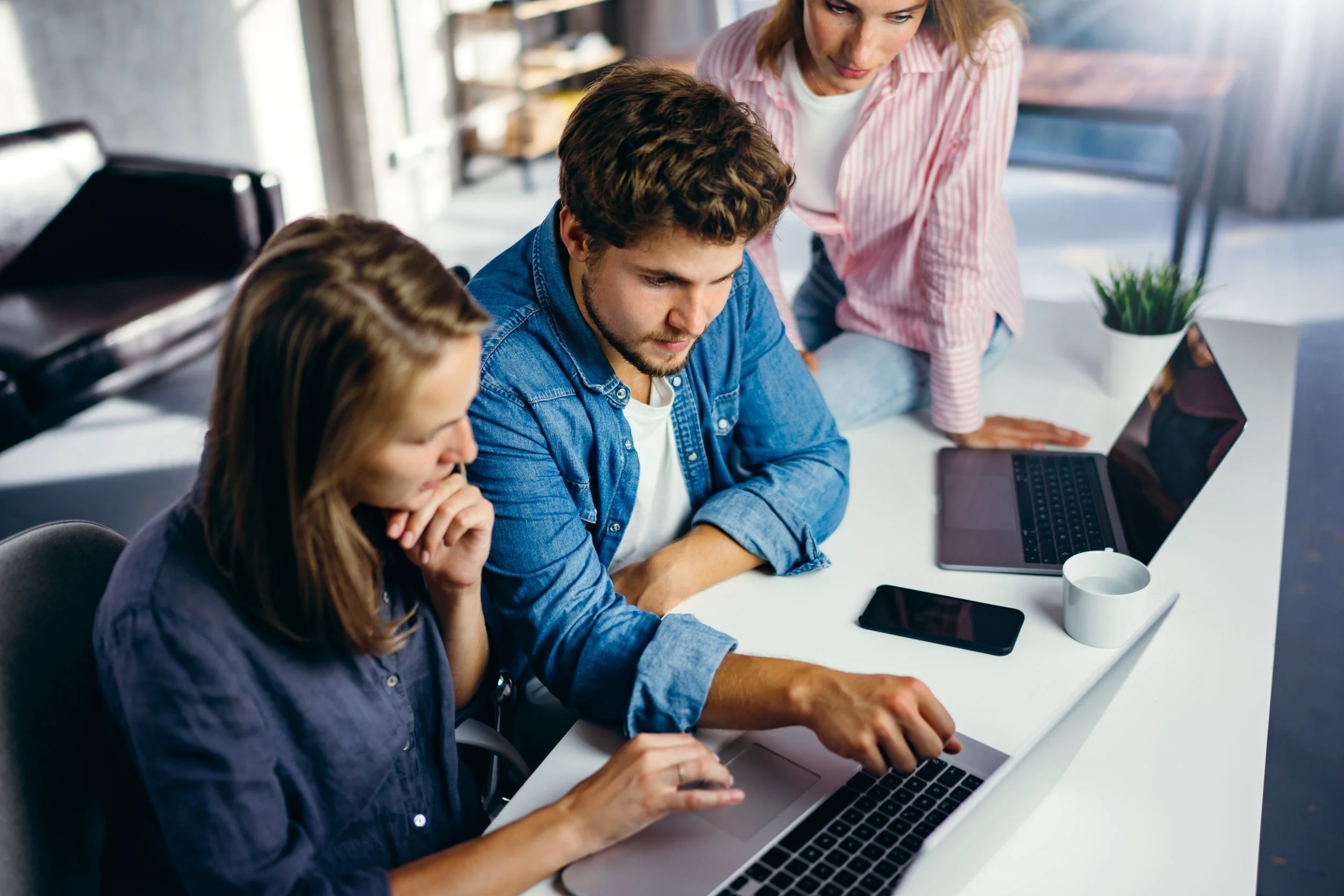 Tre personer arbejder sammen ved et bord med laptops, telefon, kop og en plante i et moderne kontormiljø.