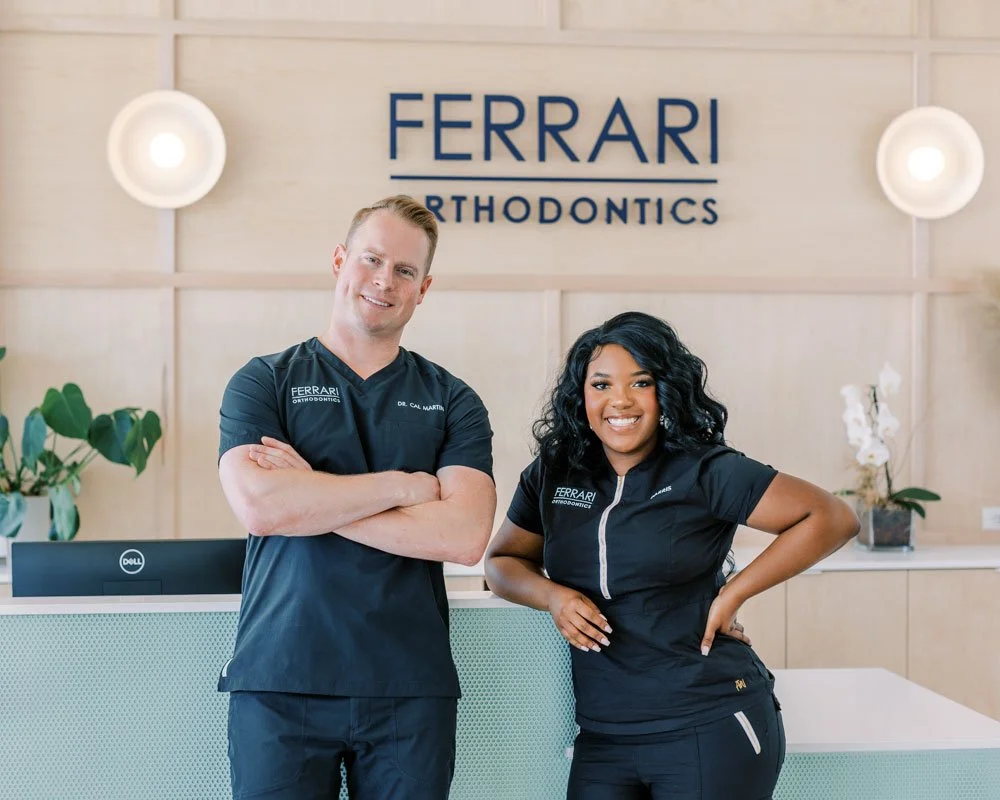 Two smiling orthodontic professionals, a man and a woman, standing inside a modern orthodontics clinic with a sign that reads 'FERRARI ORTHODONTICS' behind them.