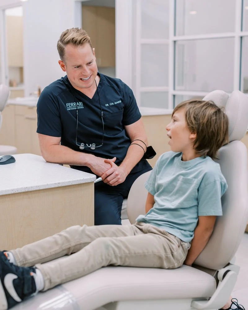 A young boy sitting in an orthodontic chair, smiling and engaging in conversation with a male orthodontist in an orthodontic office.