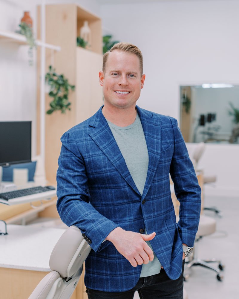 A male orthodontist in a blue blazer smiling in an orthodontic office setting with a computer and shelves in the background.
