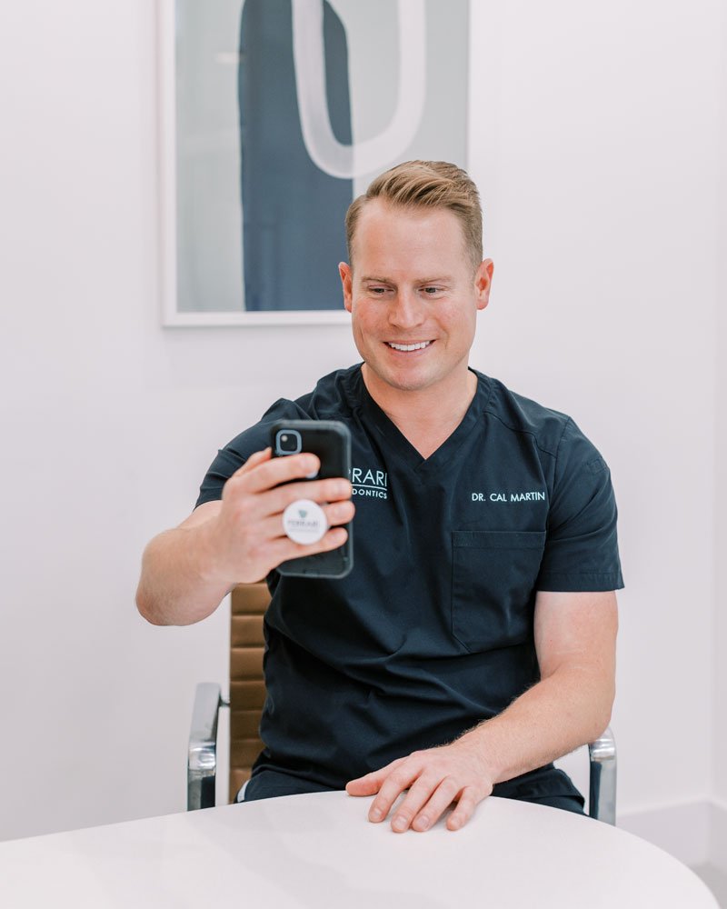 A smiling orthodontist in a dark blue medical uniform having a virtual consultation in a modern clinic setting.
