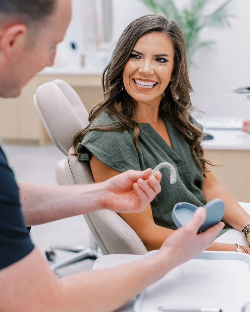 A woman smiling at an orthodontist at an orthodontic office, holding a clear aligner and a plastic case.