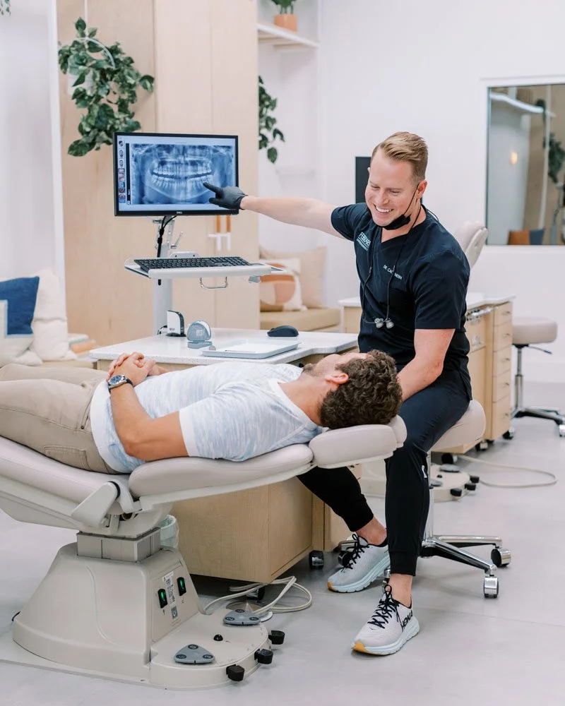 An orthodontist showing a digital dental X-ray of a patient's teeth on a monitor in a dental office, with the patient lying on the dental chair.