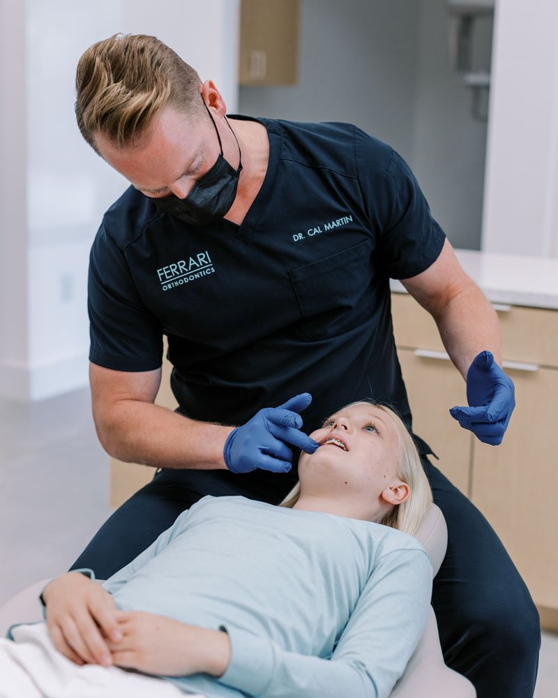Orthodontist in black scrubs and face mask examining a young girl in a dental chair with a dental mirror.
