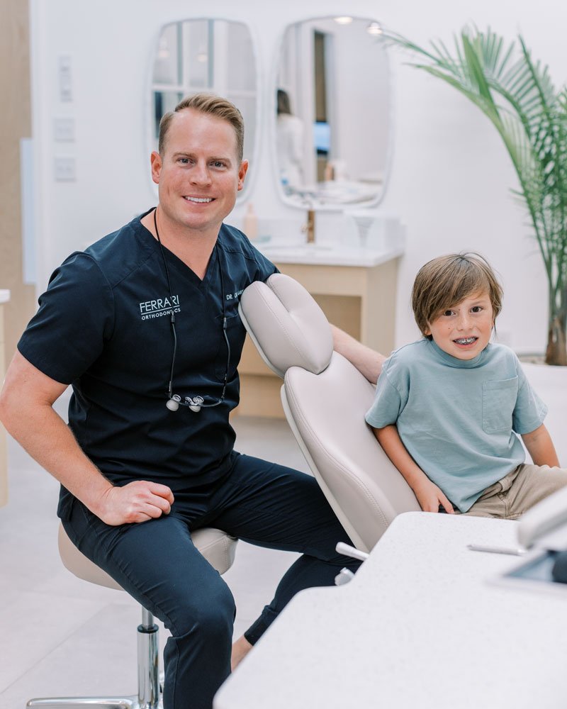 A smiling male orthodontist in navy scrubs sitting next to a young boy with brown hair wearing a light blue shirt, seated in a dental chair in a modern orthodontic office.