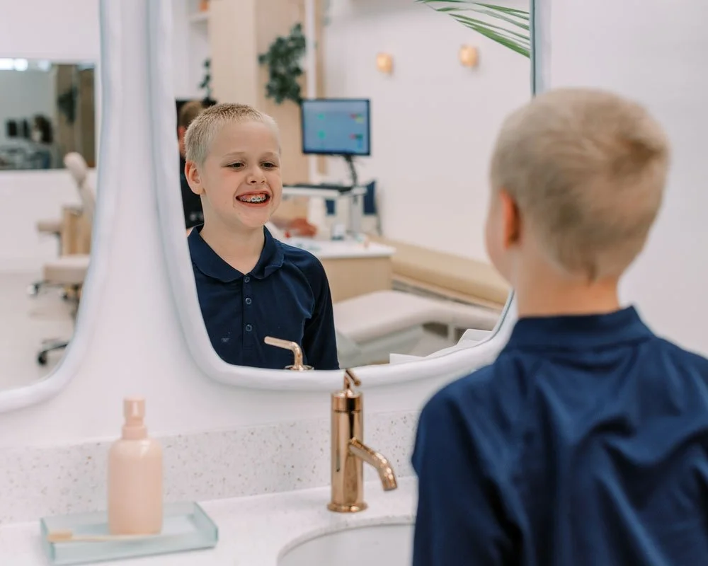 A young boy with braces looking at himself in a mirror in a modern clinic, with a soap dispenser and a sink in the foreground.