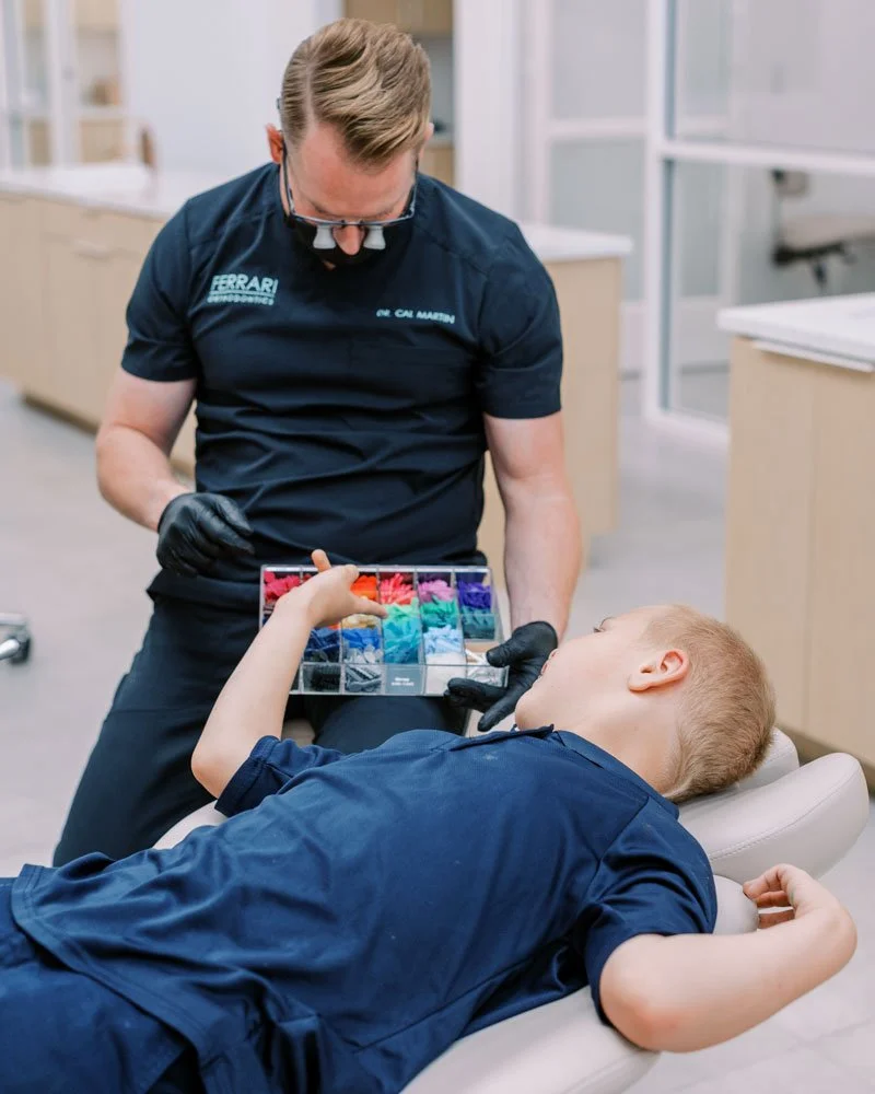 An orthodontist wearing magnifying glasses and gloves shows a tray of colorful dental floss to a young boy lying in a dental chair.
