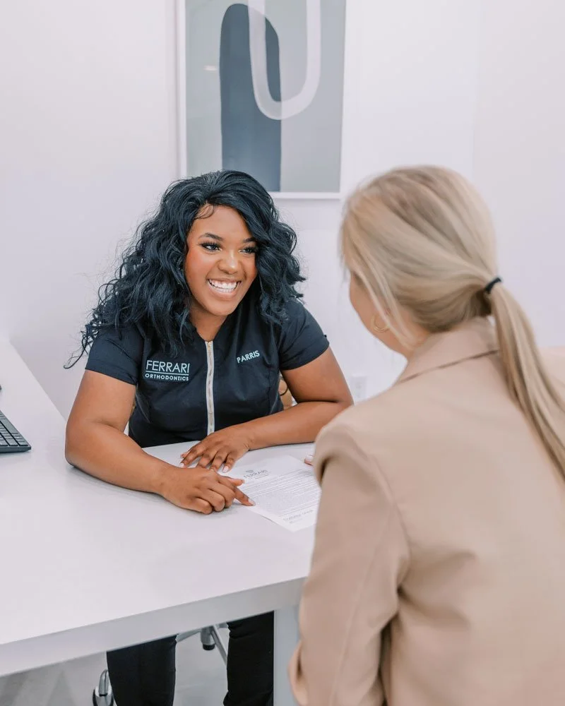 A woman with curly black hair smiling and talking with a woman with blonde hair tied back, sitting at a white desk in an orthodontic office.