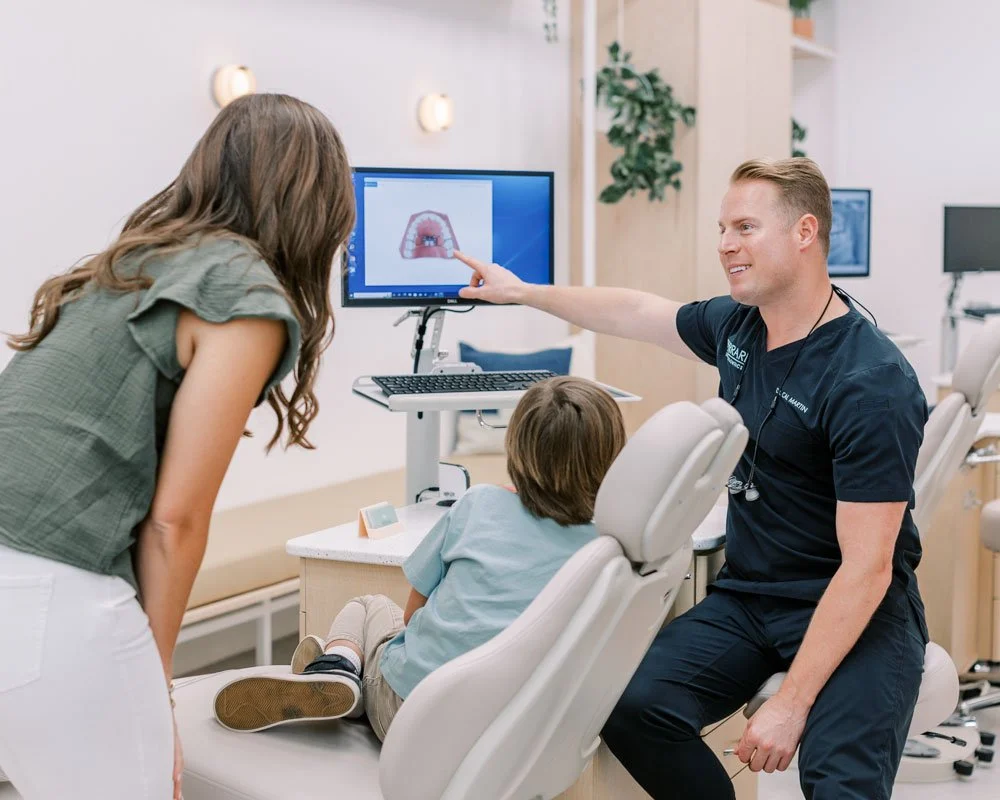 An orthodontist shows a child a diagram of a mouth on a computer monitor during a dental consultation, with the child's mother standing nearby.