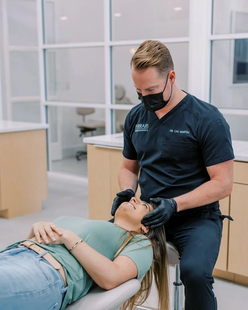 An orthodontist in scrubs and a face mask examines a patient lying on a dental chair.