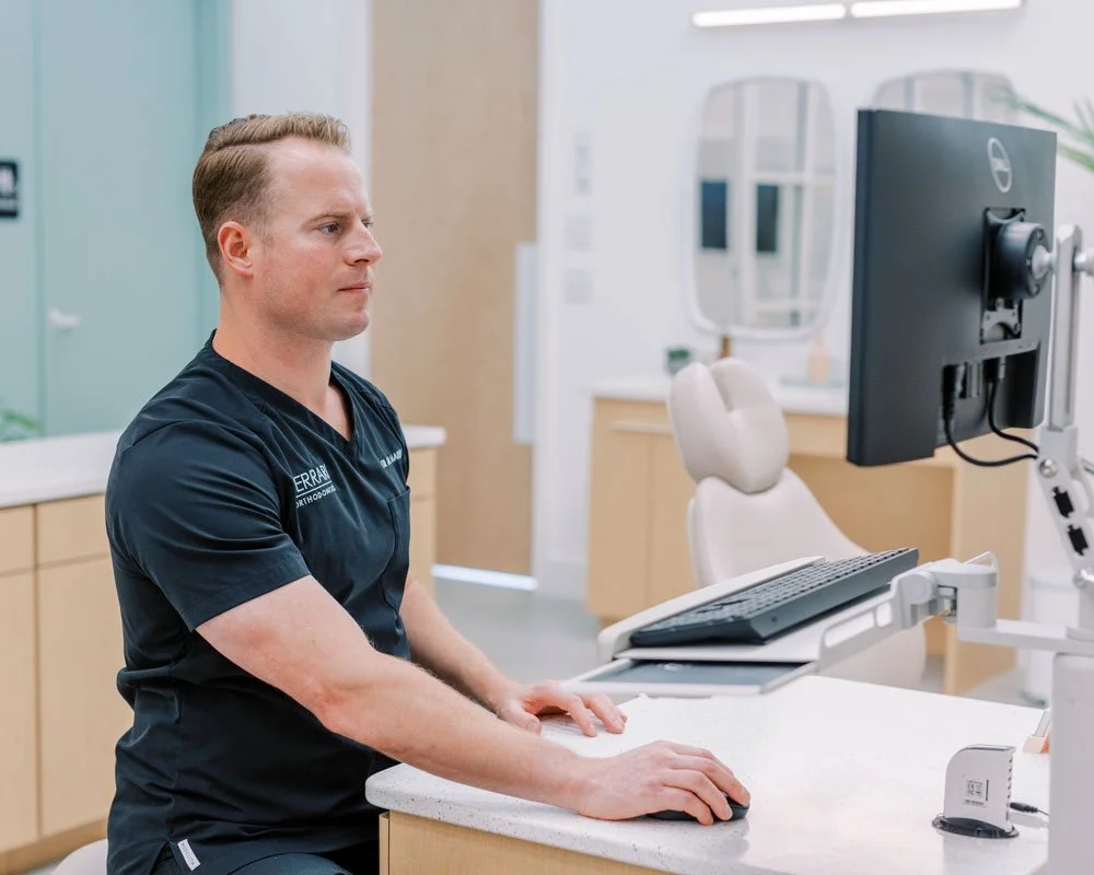 An orthodontist in a navy scrubs shirt working at a computer in a dental clinic.