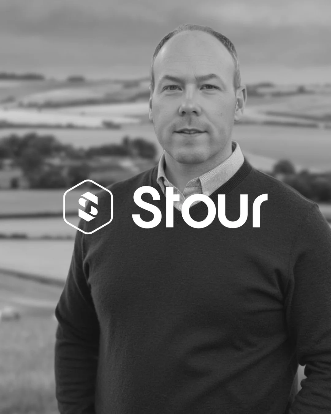 Black and white photo of a man standing outdoors in front of fields with a cloudy sky. The man is wearing a collared shirt under a sweater. The logo and text 'stour' are overlaid on the image.