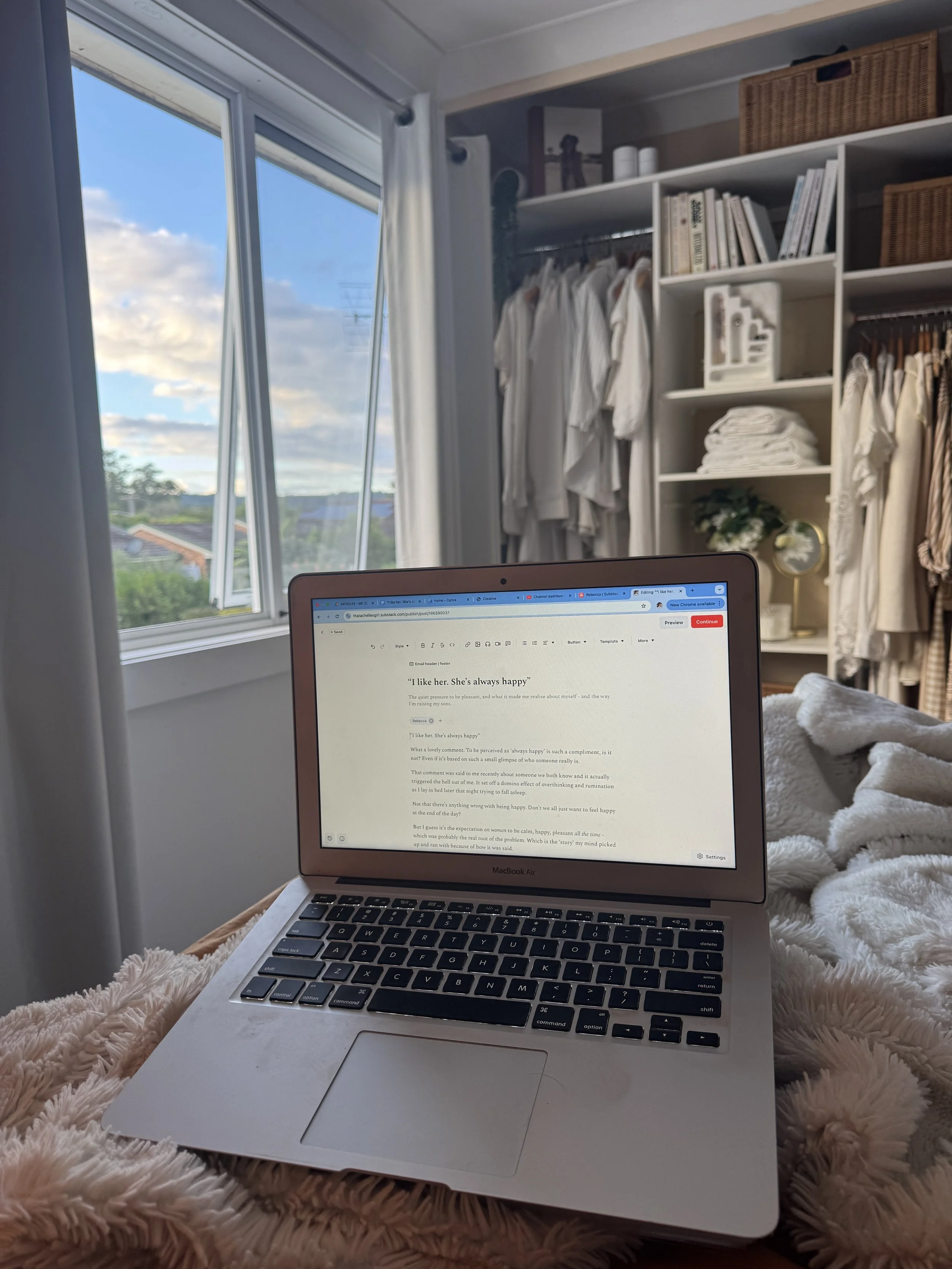 Rebecca (Achelles) Smith, writing. A laptop on a plush, cream-colored blanket, with a background of open window showing a partly cloudy sky and rooftops.