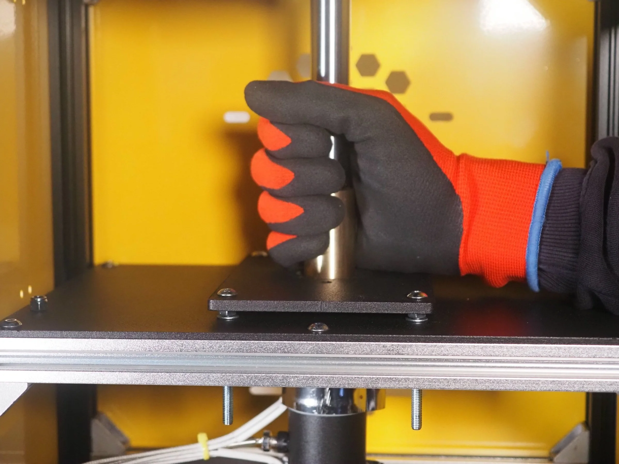 A person wearing a black glove with red fingertips is adjusting a metal component inside a yellow and black 3D printer or machinery.