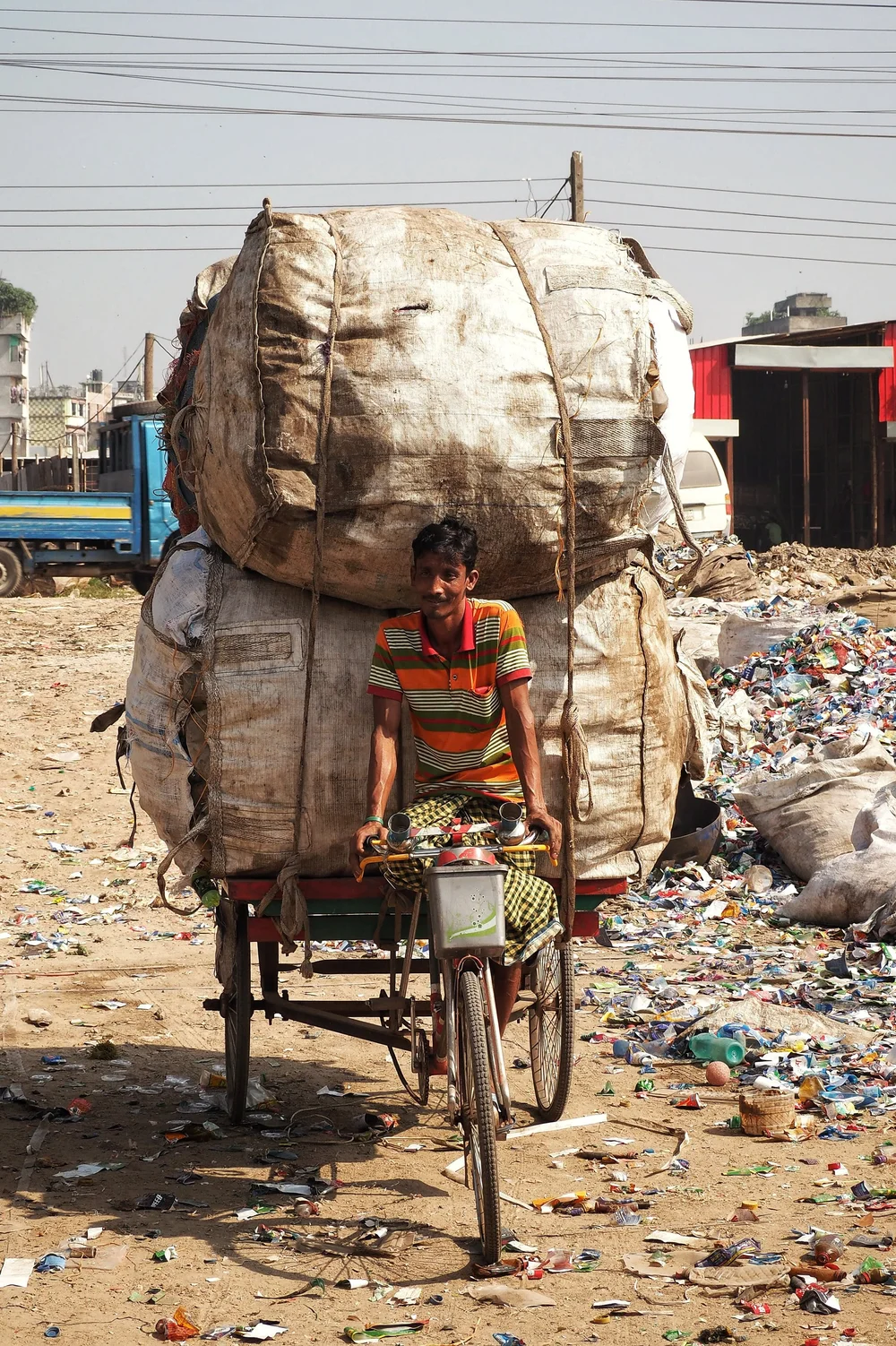A waste picker in Bangladesh carries a bag of collected plastic to a buying station, paid per kilogram on the spot.