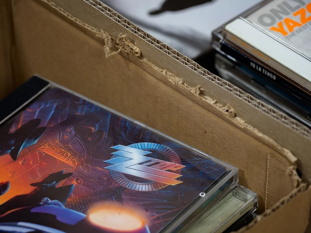CD and cassette cases stacked in an open cardboard box.