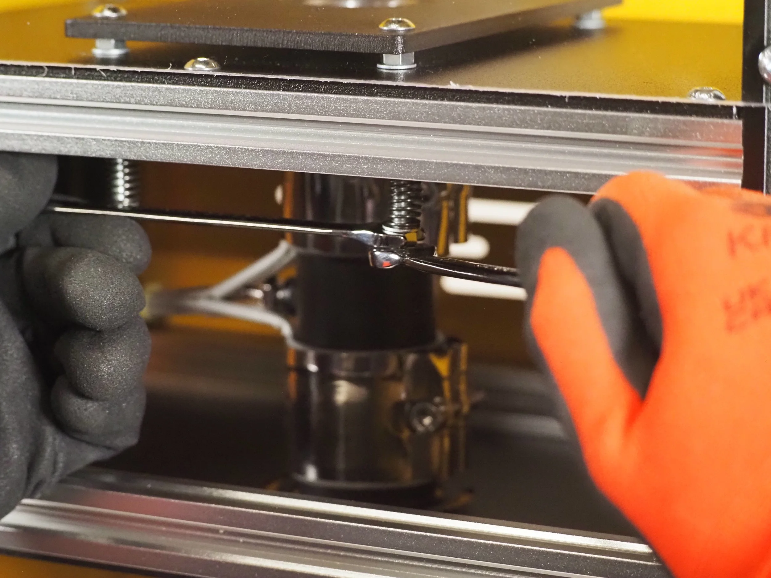 Close-up of a person wearing gray and orange gloves, assembling or repairing a mechanical device, possibly a 3D printer or CNC machine, with metal parts, screws, and a frame.