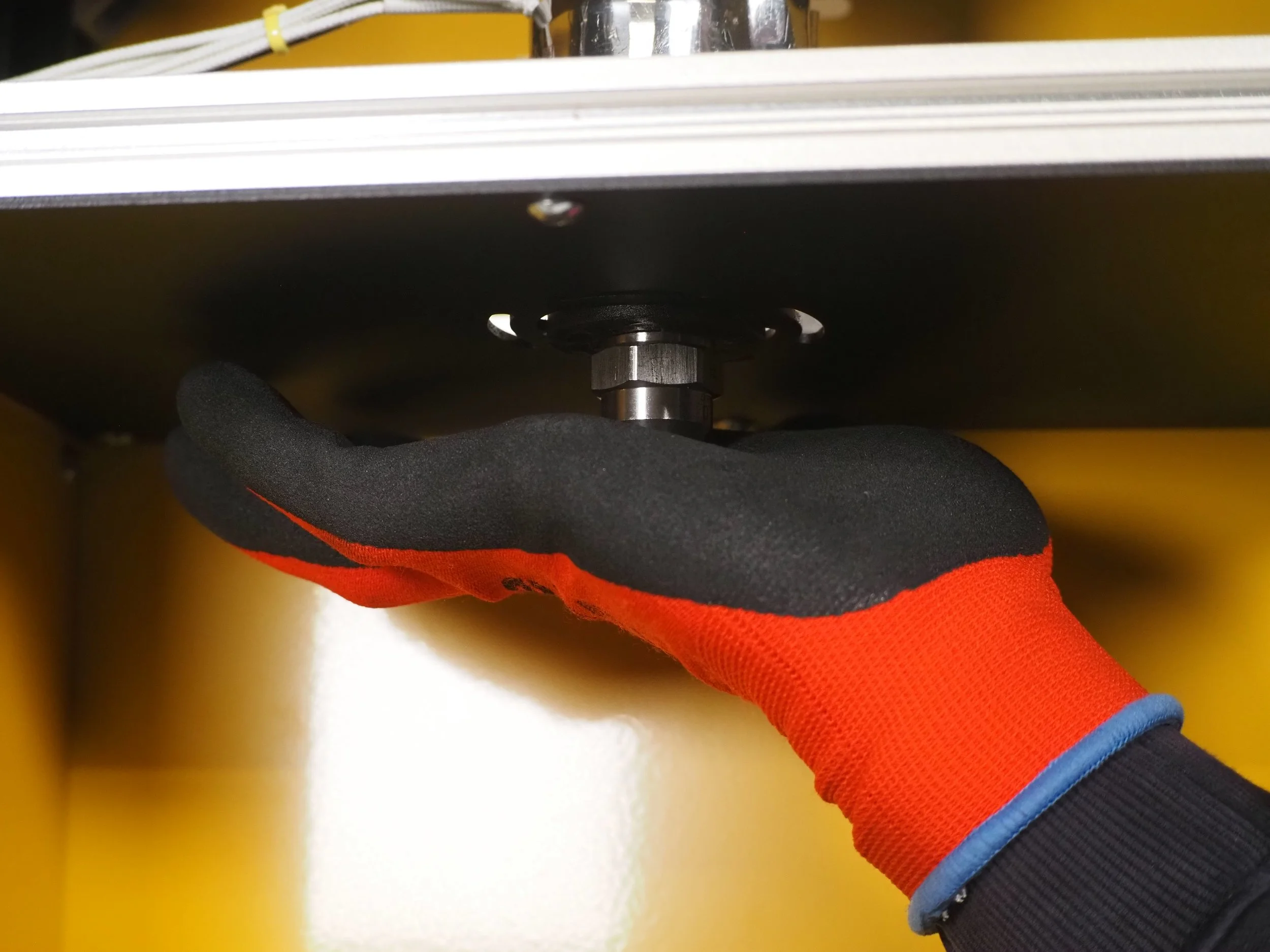 A person's gloved hand is adjusting or holding a mechanical component under a yellow surface, possibly part of machinery or equipment.