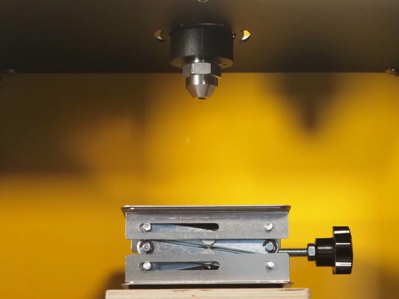 Close-up of a CNC machine or milling device with a metallic cutting tool, mounted on a dark gray surface with a yellow background.
