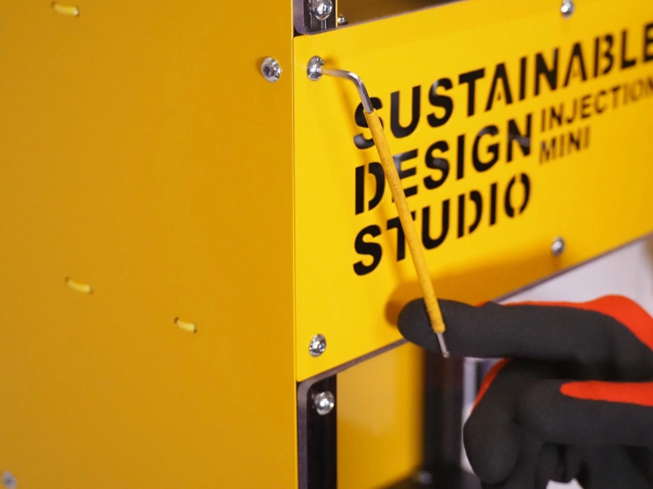 A person wearing gloves using a tool to install a yellow sign that reads 'SUSTAINABLE DESIGN MINI STUDIO' on a yellow metal box.