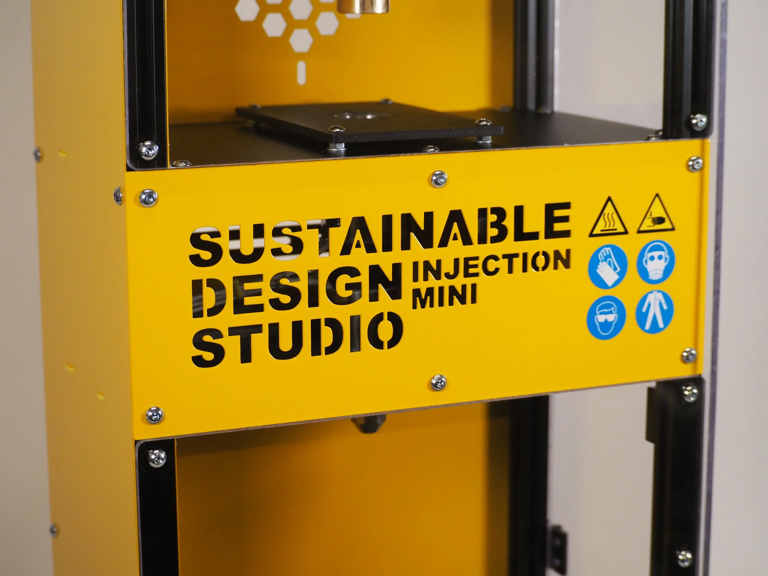 Yellow 3D printer with a label reading 'Sustainable Design Injection Mini Studio' and safety icons for heat and hot surface, hand protection, mask, goggles, and protective clothing.