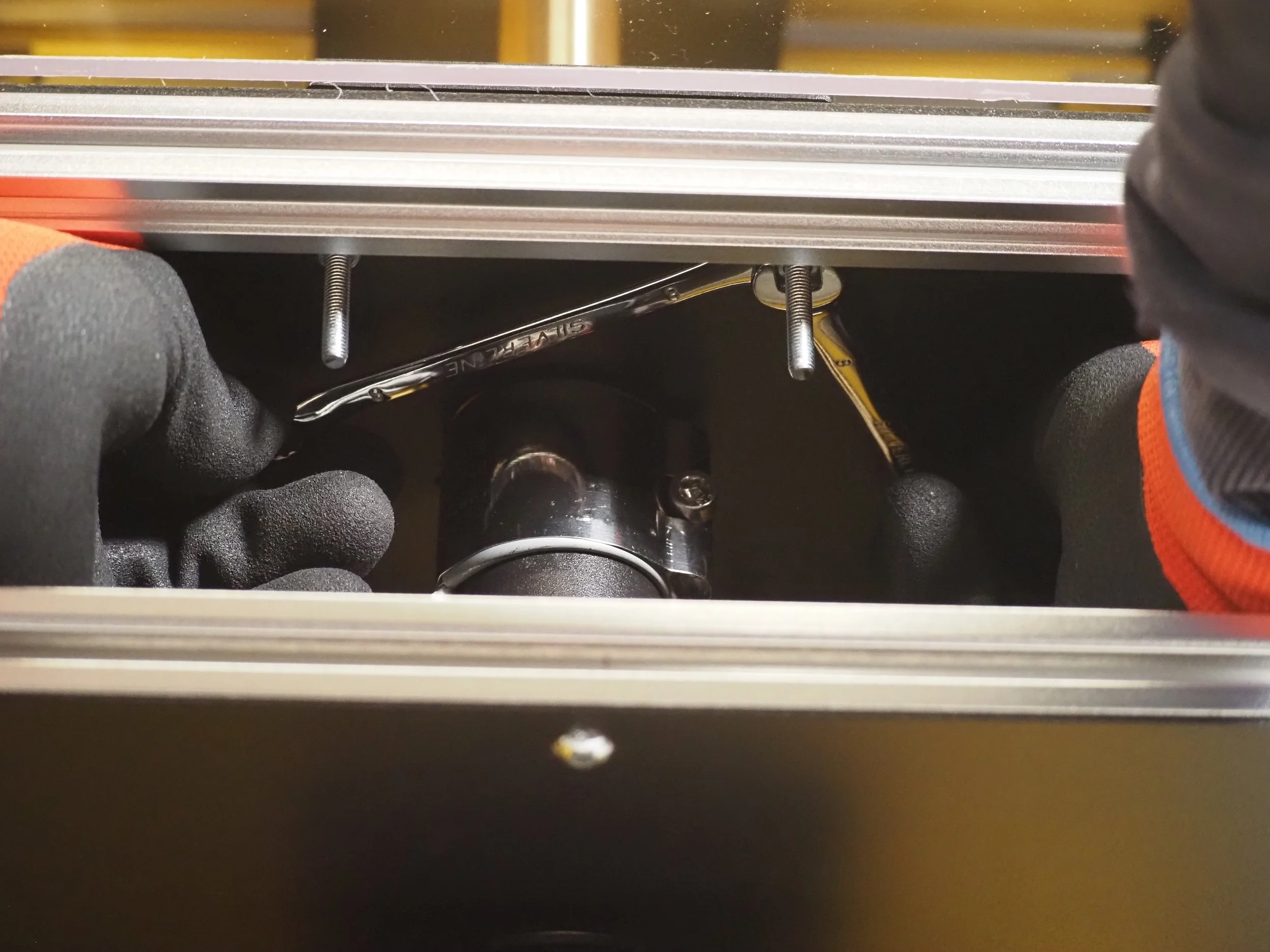 Close-up of a mechanical component inside a metal frame, with black gloves and hands, including a steering linkage and bolts.