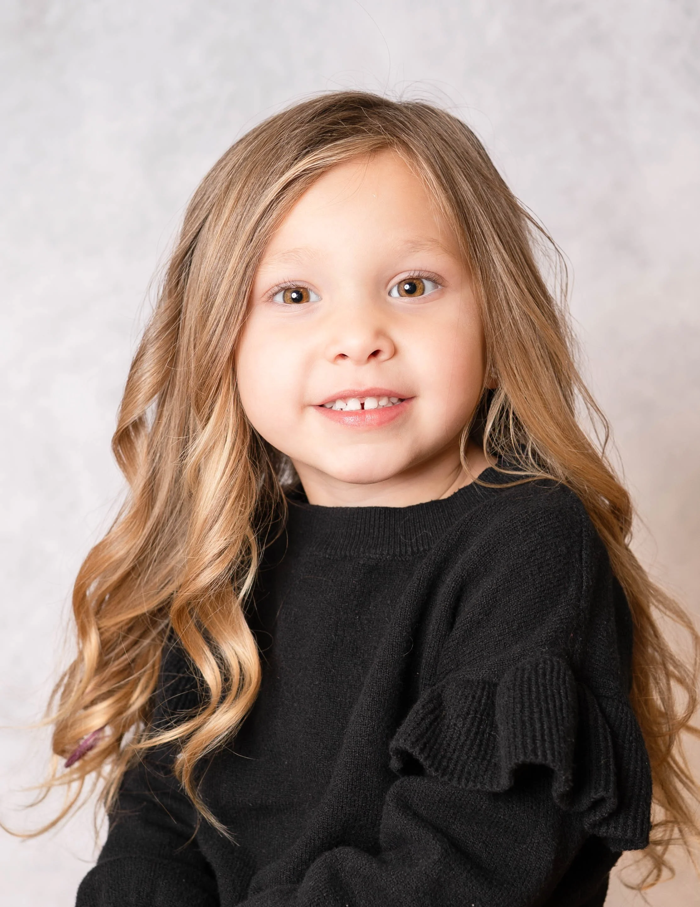 Close-up of a young girl with long, wavy blonde hair, light skin, and a big smile showing some missing front teeth, wearing a black sweater, against a plain light background.