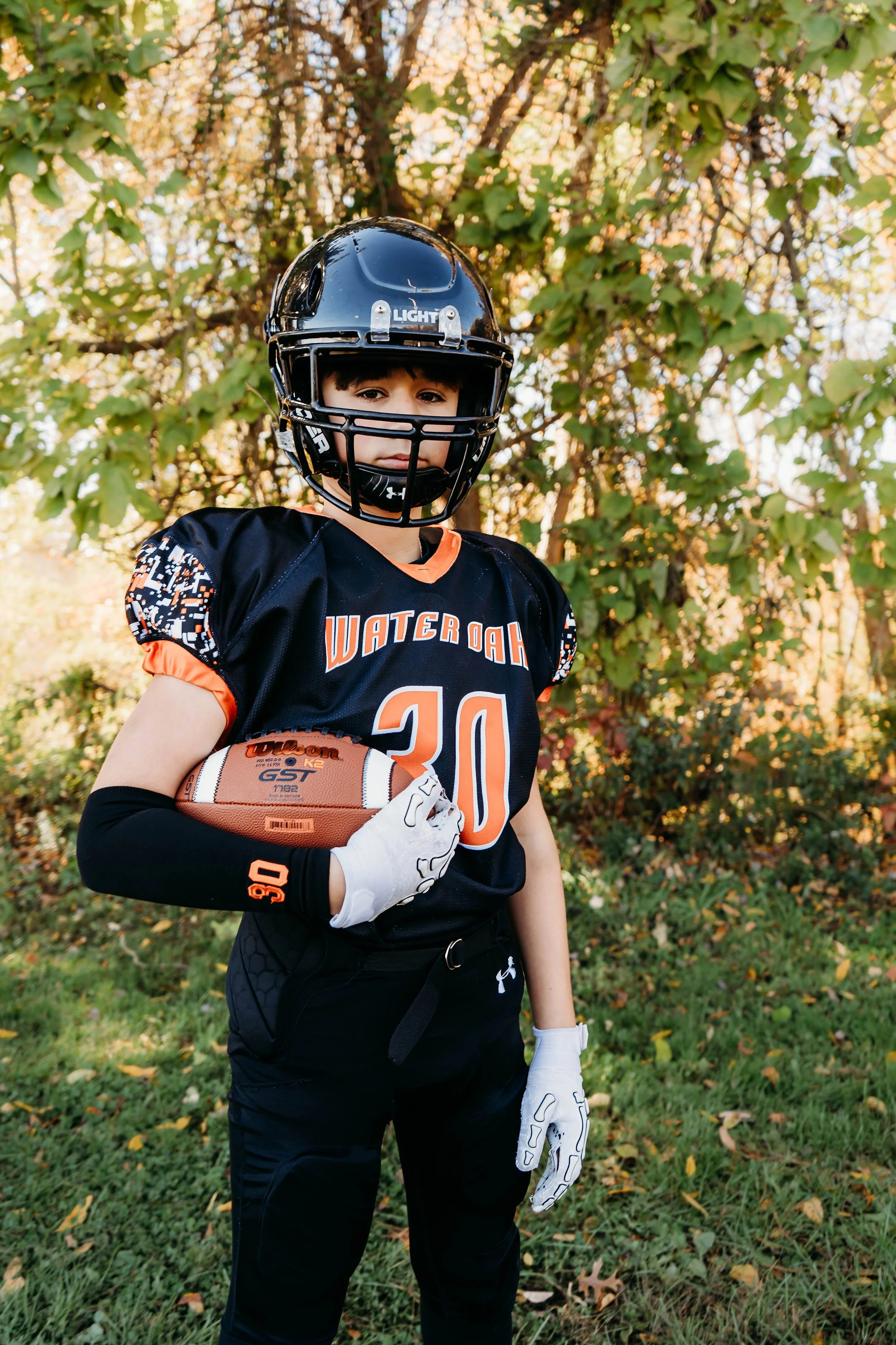 A young boy wearing American football gear, including a black helmet and black jersey with orange and white accents, holding a football and standing outdoors in a park with autumn foliage in the background.