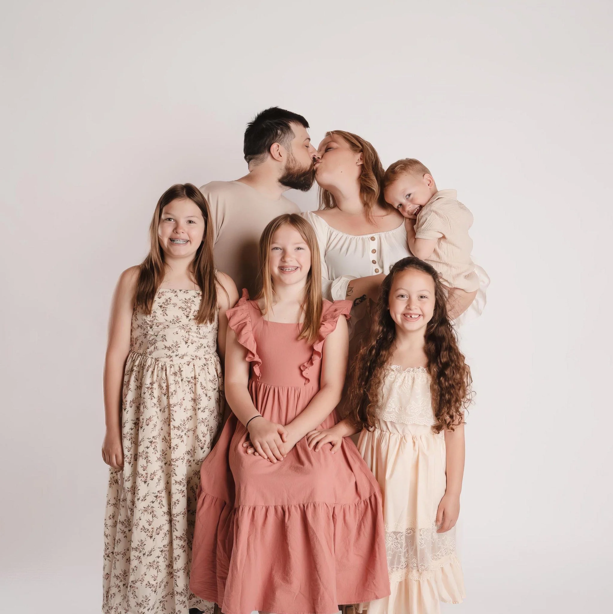 A group of children and adults, including a man and woman, standing in front of a plain white background. The woman is kissing the man on the lips, while the children smile at the camera.