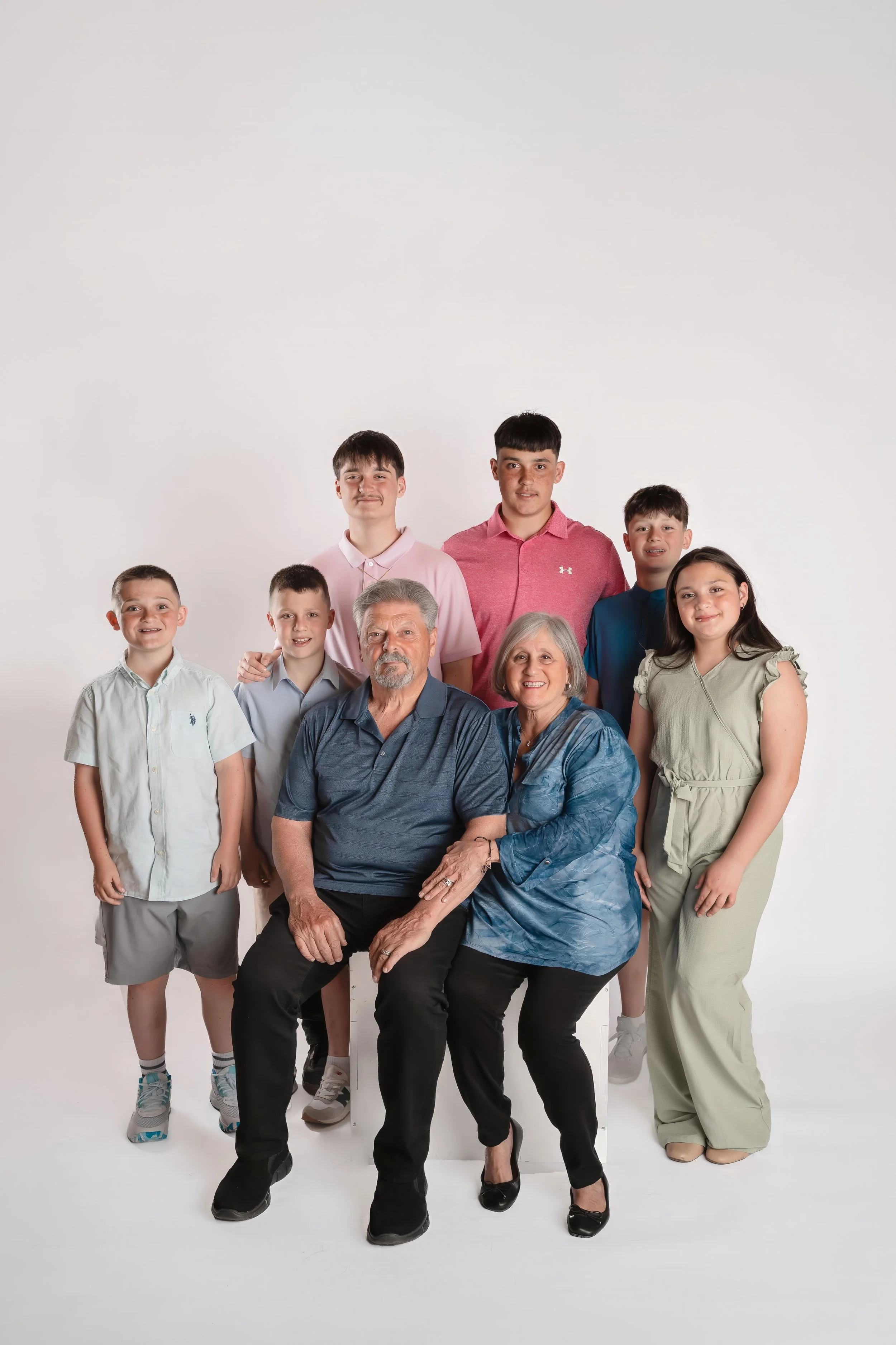 A multigenerational family photo with three elderly adults seated in the front, surrounded by five children and teenagers standing behind them, all smiling against a plain white background.