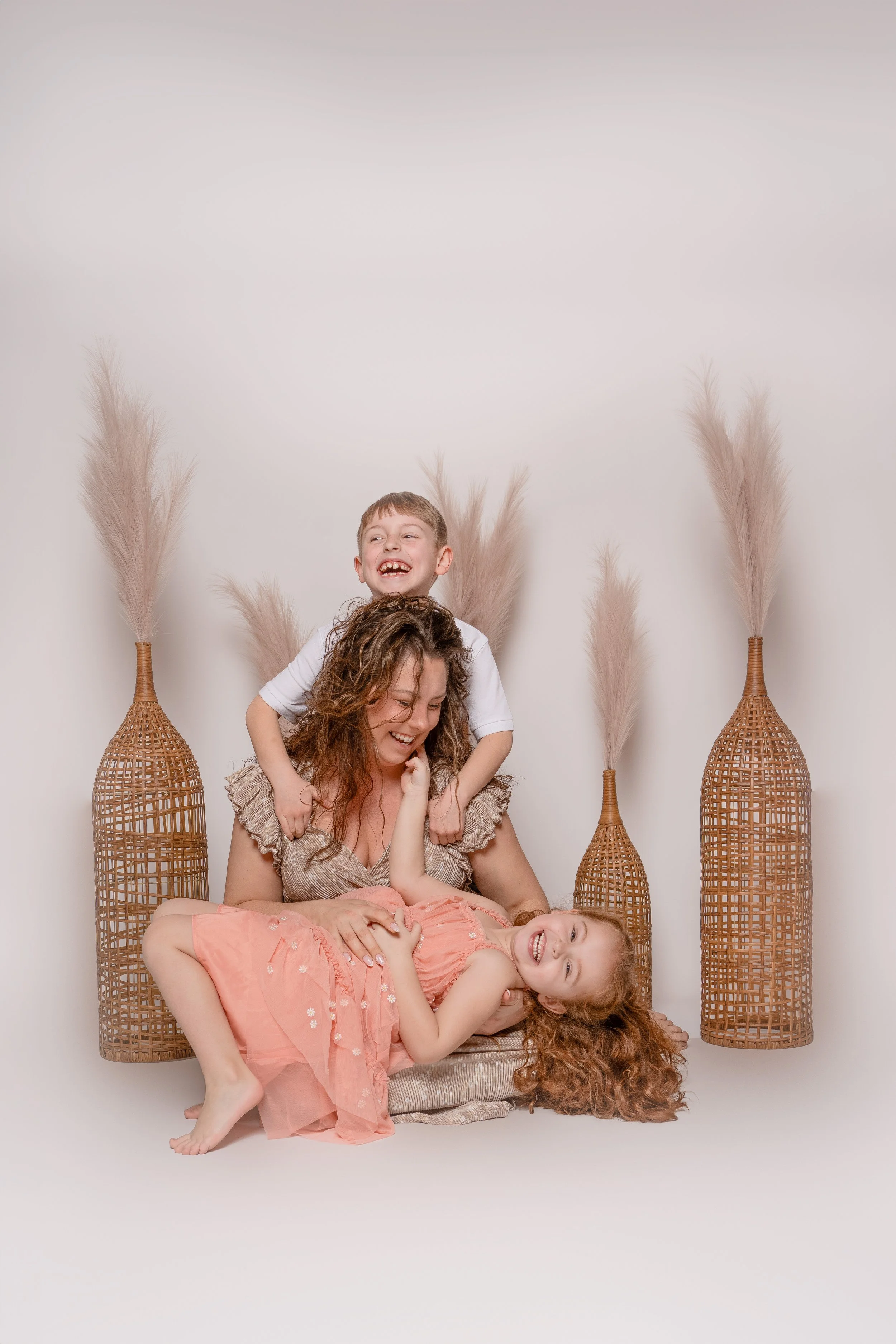 Four children, three girls and one boy, playing together and smiling in front of a plain white background with tall, woven vases with pampas grass.