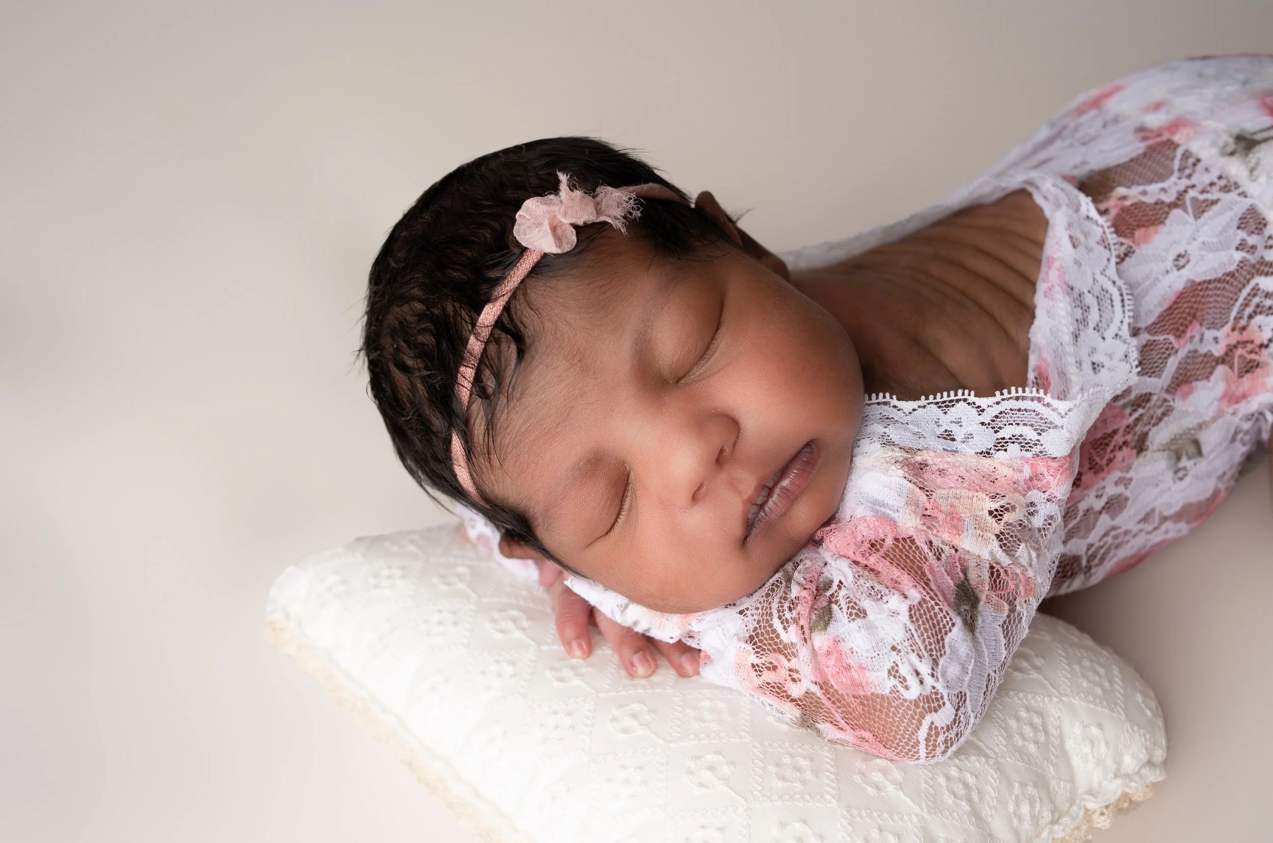 Close-up of a sleeping infant with dark hair, wearing a pink headband with a bow, dressed in a pink and white lace dress, resting on a white textured blanket against a plain light-colored background.