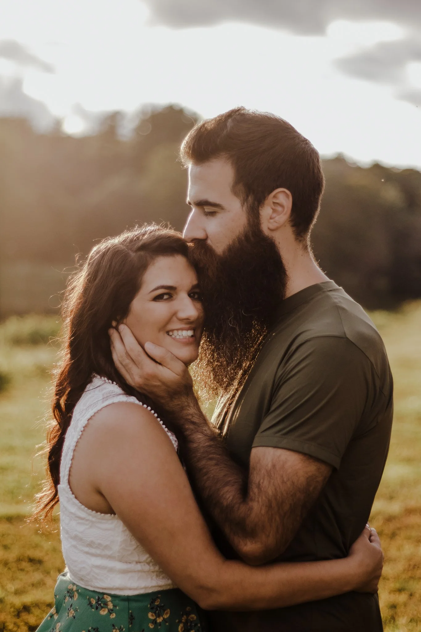 A couple stands close together in a field with trees and a partly cloudy sky in the background. The woman has long, wavy brown hair, is smiling, and is wearing a white top and a green skirt with floral patterns. The man has a full beard, brown hair, and is wearing a dark t-shirt. They are embracing each other affectionately.