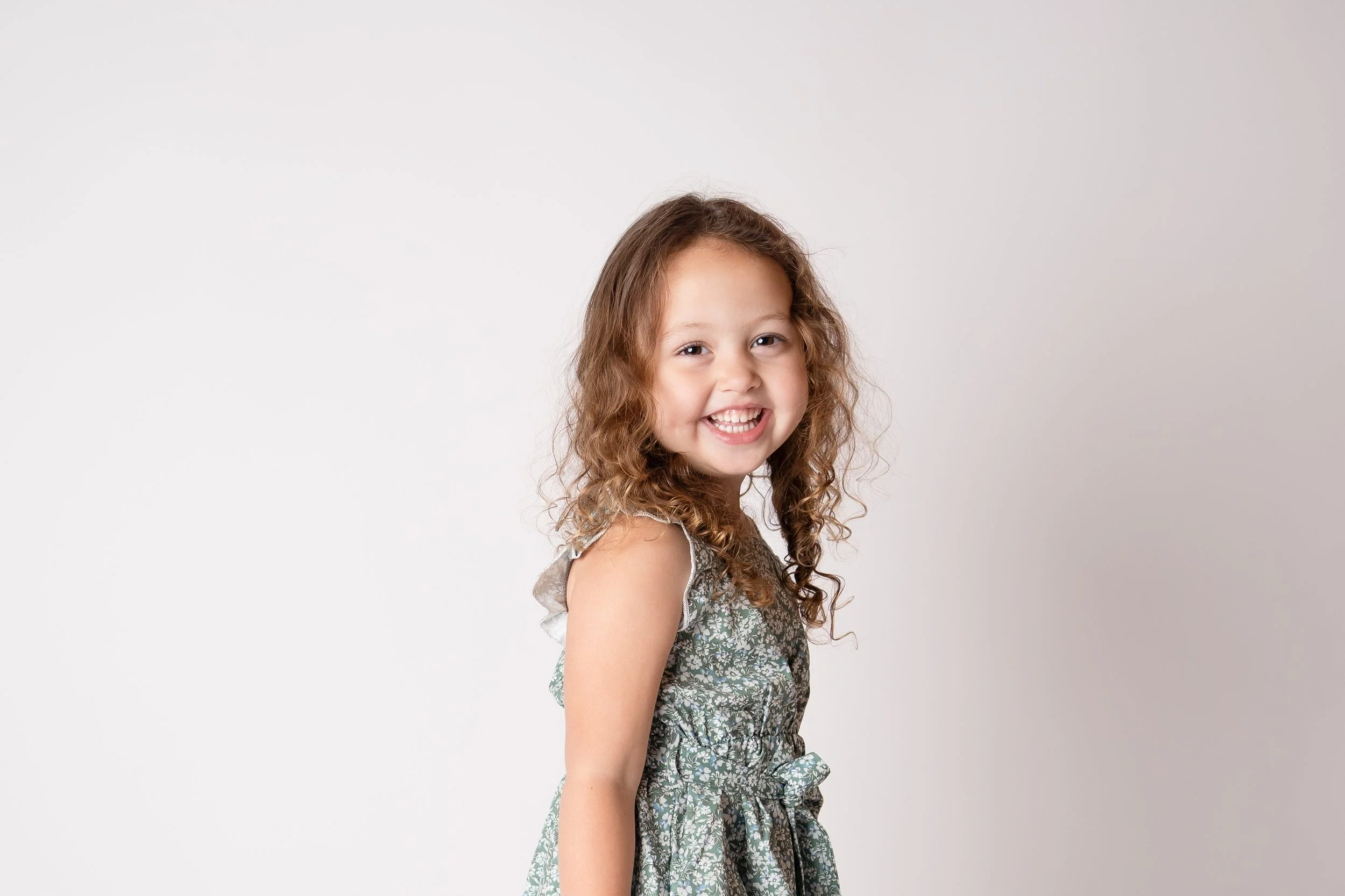 A young girl with curly brown hair smiling at the camera, wearing a floral dress, standing against a plain white background.