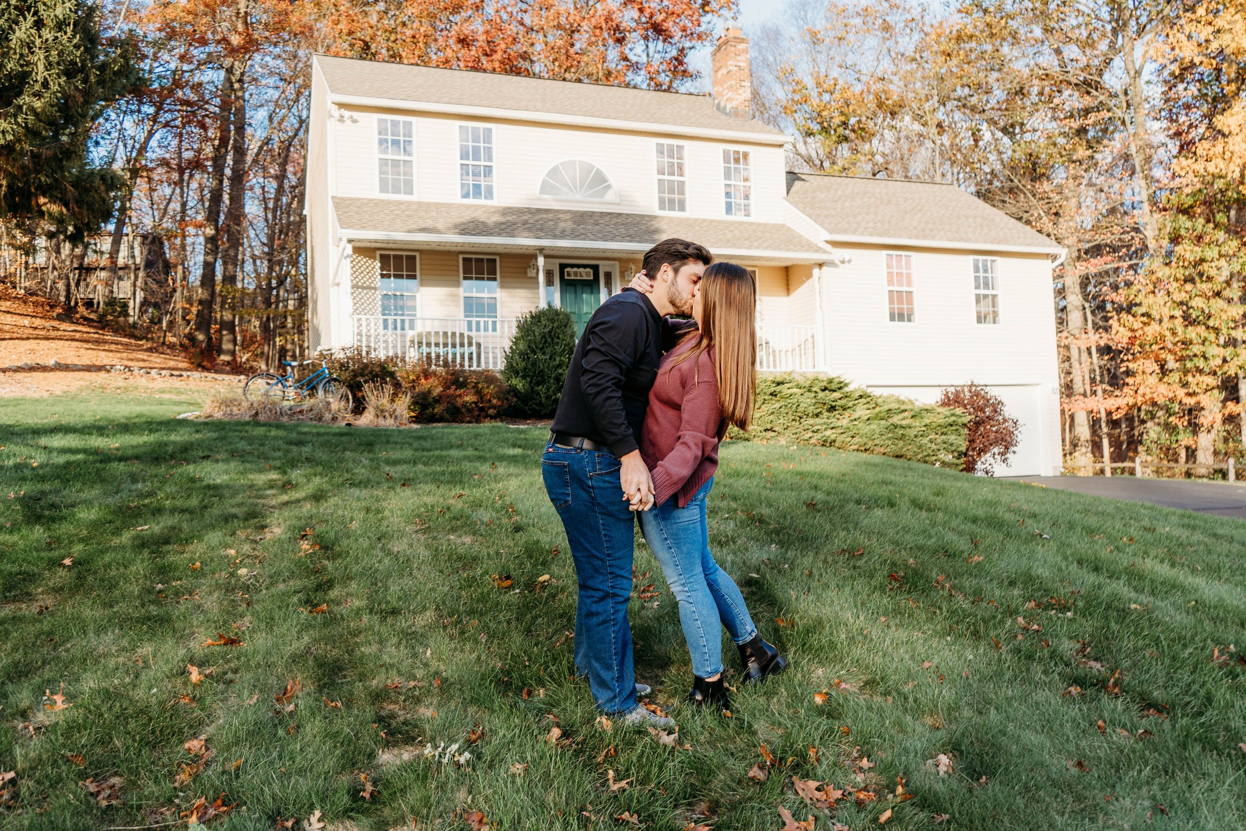 A couple kissing in front of a white house with large windows and a porch. The yard has green grass and fallen autumn leaves, with trees showing fall foliage in the background.