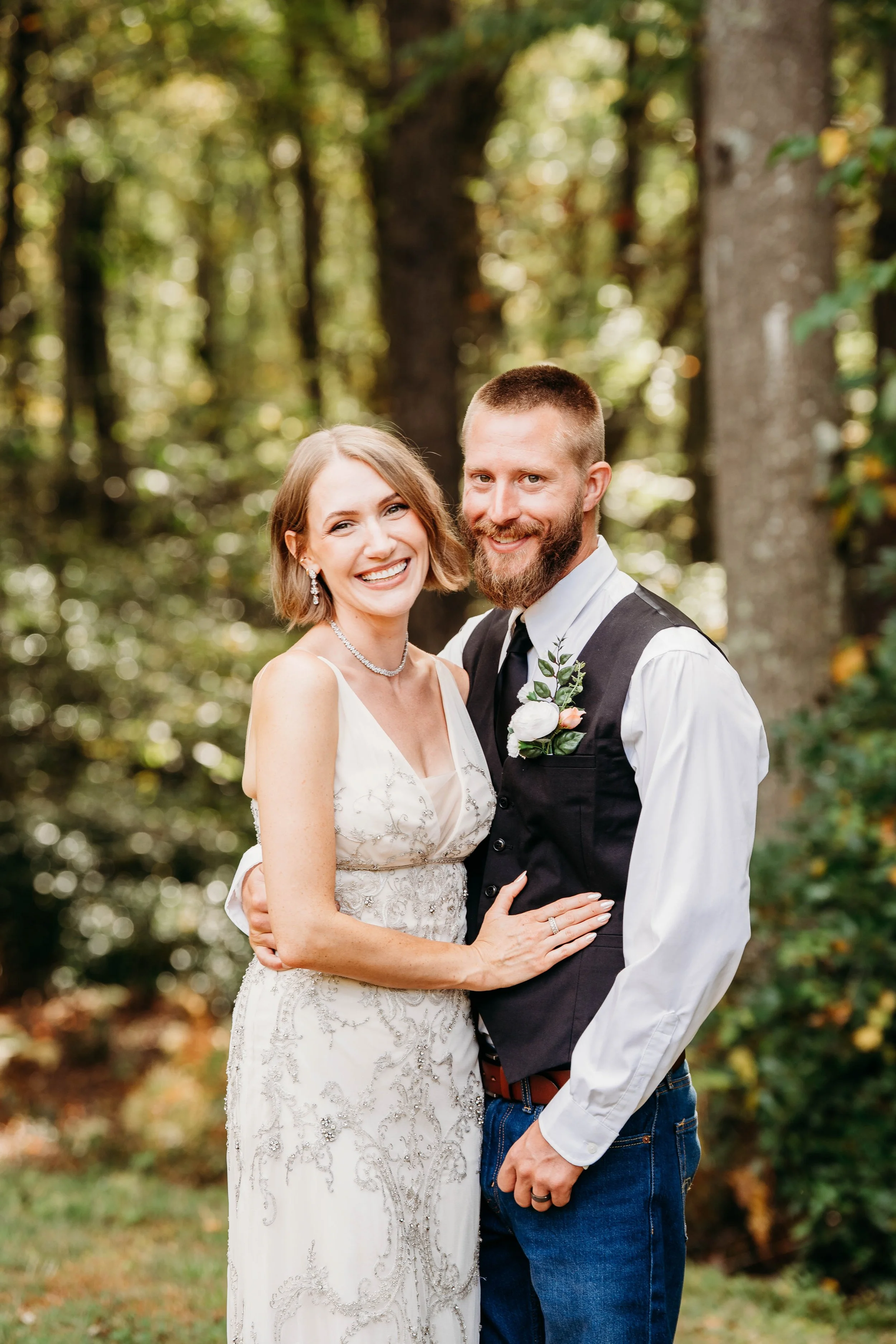 A smiling bride and groom in wedding attire standing outdoors in a forest setting with tall trees and green foliage in the background.