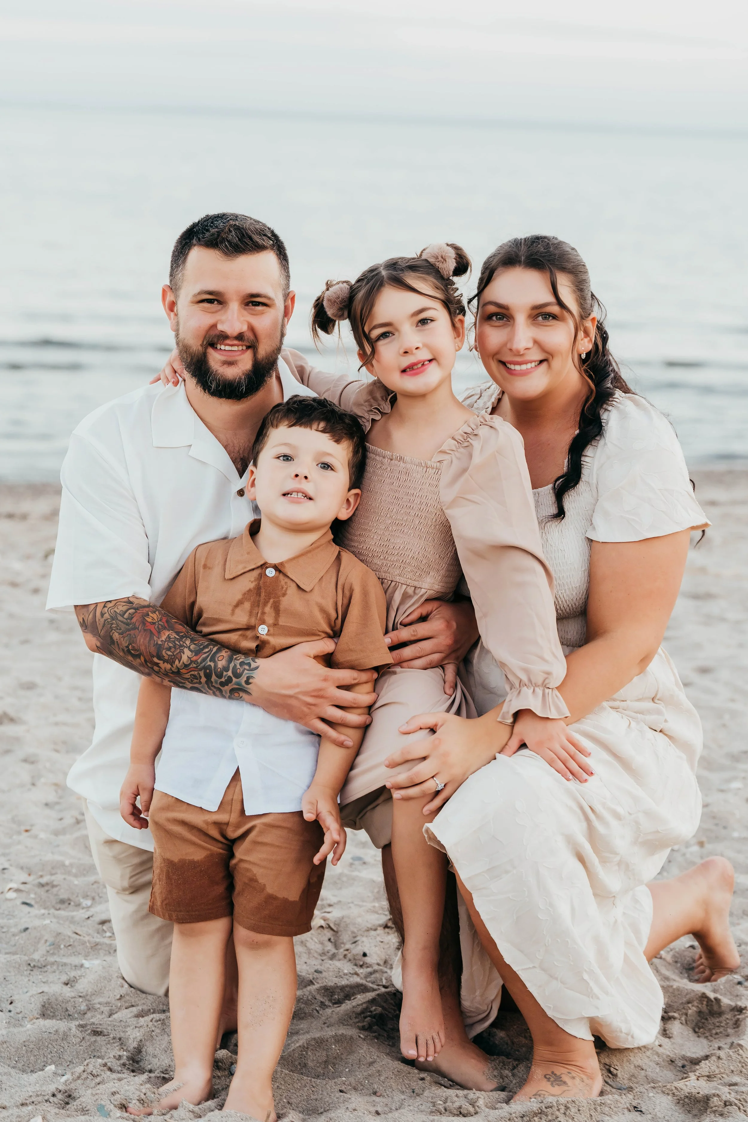 A happy family of five on the beach with the ocean in the background. The father has a beard and tattoos on his arm, wearing a white shirt. The mother has long dark hair and is dressed in a cream-colored dress. The daughter has her hair in two buns with fur hair accessories, and is wearing a beige dress. The two young boys are in front, one with dark hair and a brown shirt, and the other with short hair and a beige shirt.