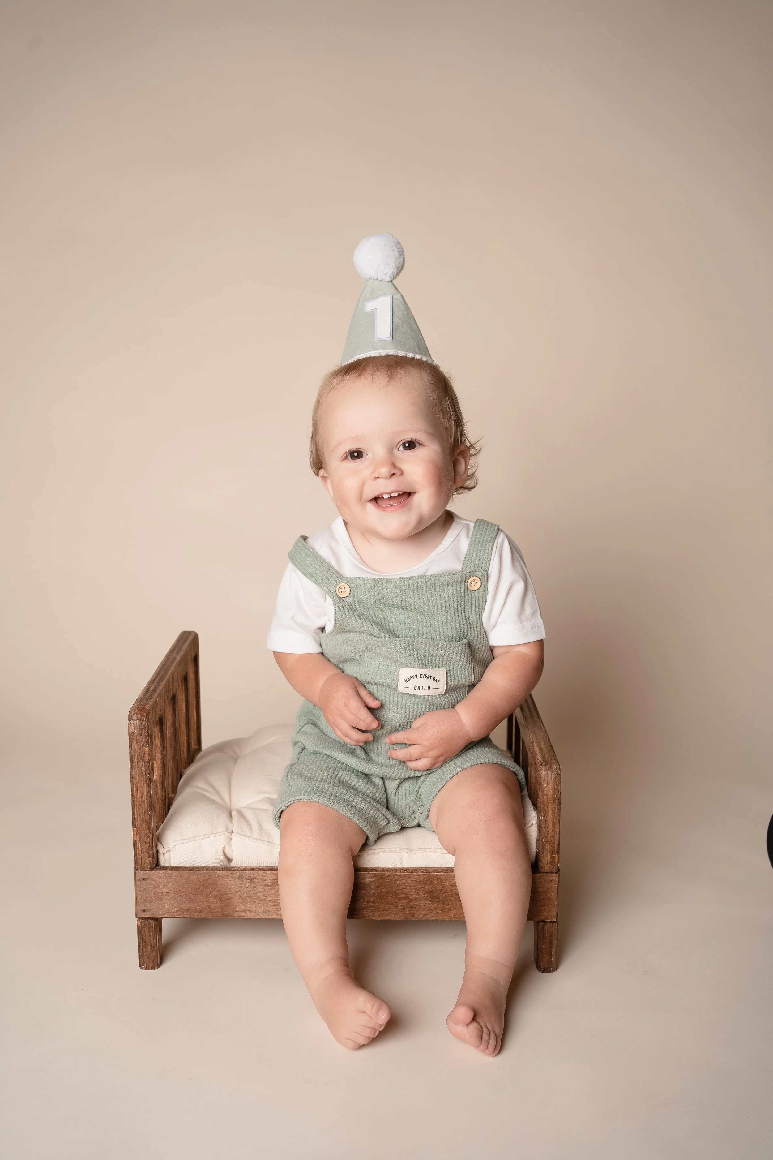 A smiling toddler wearing a party hat with the number 1, sitting on a small wooden chair with a cream cushion, against a plain beige background.