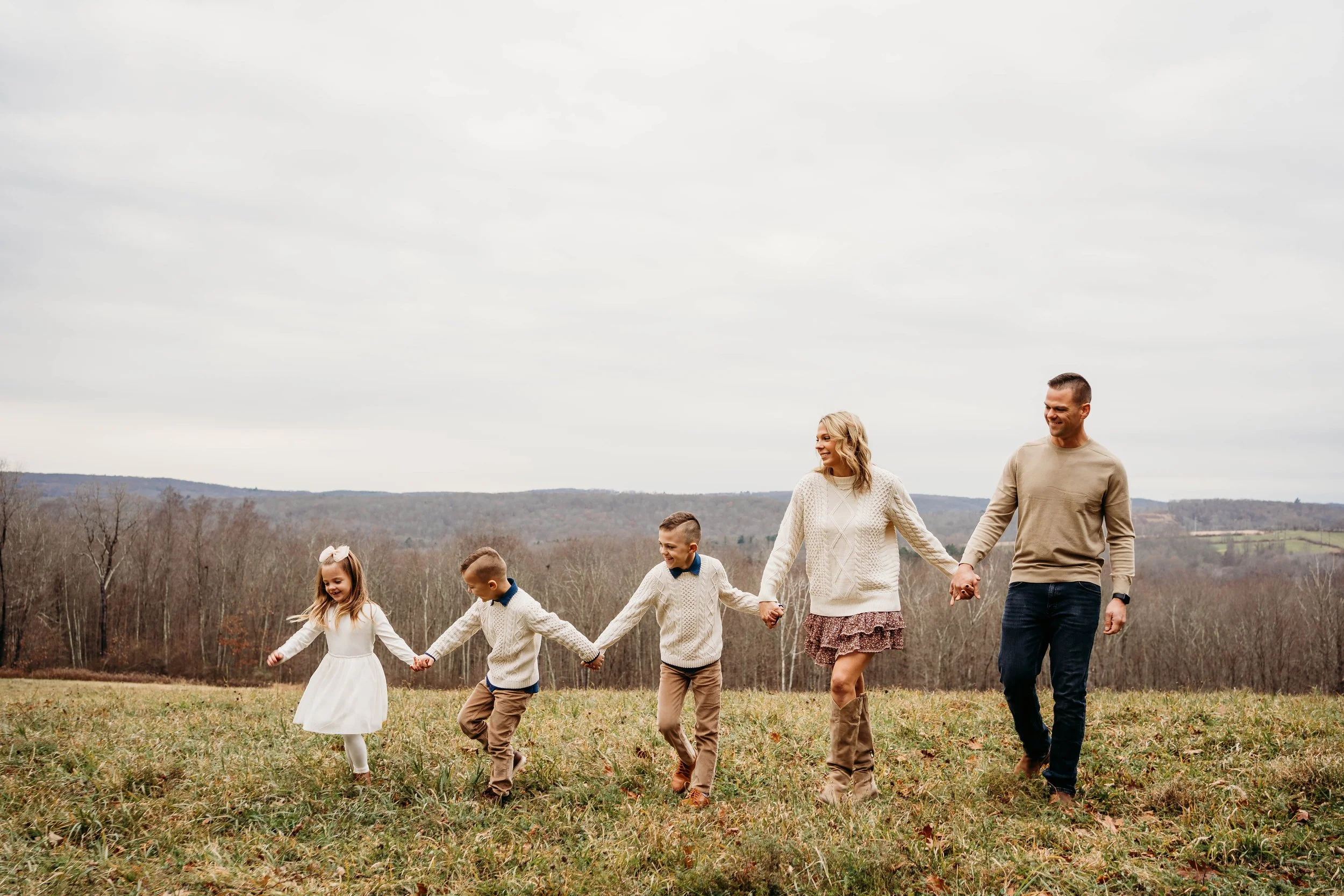 A family of six holding hands and walking together outdoors in a grassy field, with leafless trees and hills in the background under a cloudy sky.