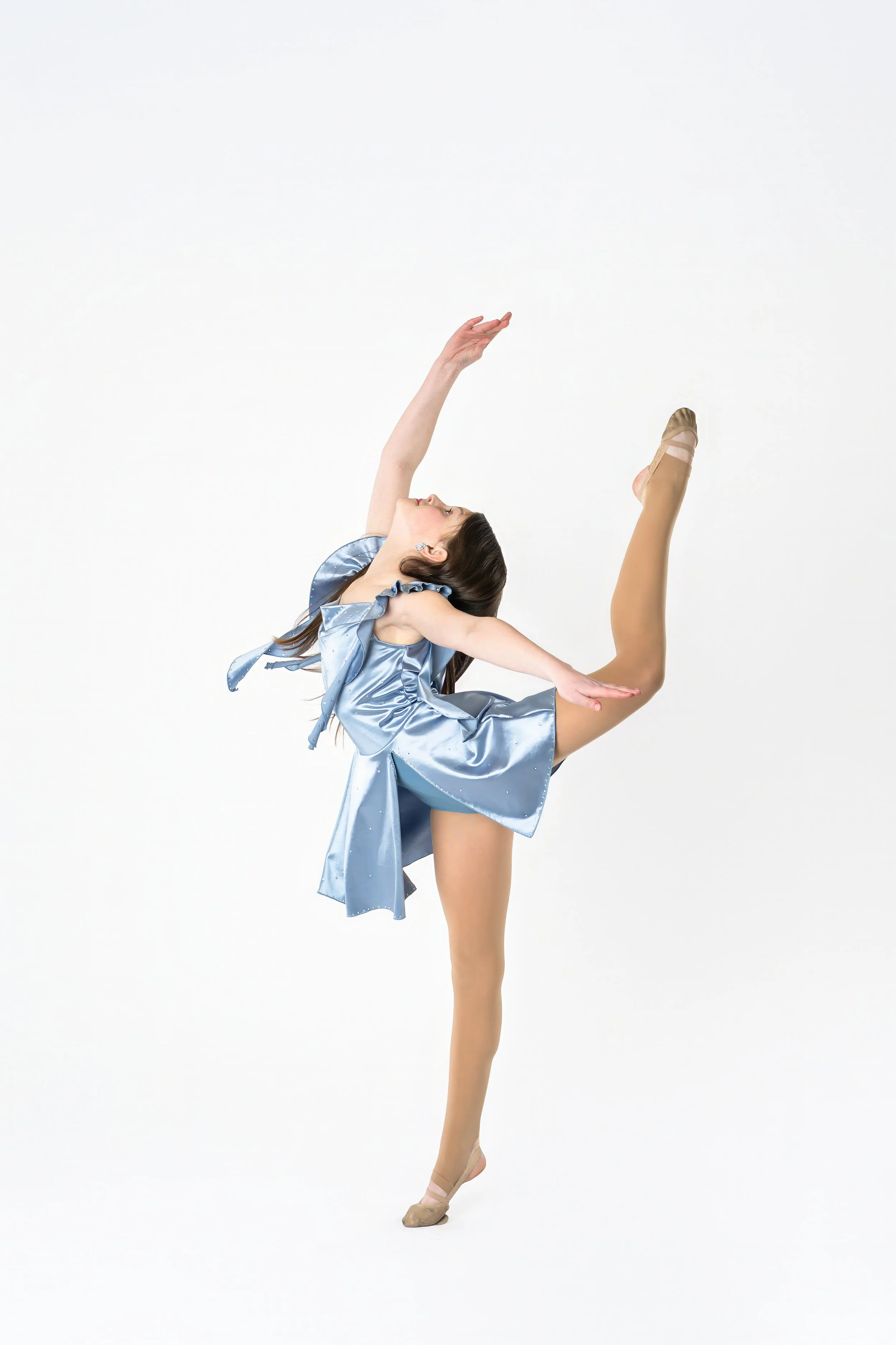 A ballerina wearing a shiny blue dress and beige ballet shoes, performing a pose with one leg raised and bent at the knee, against a plain white background.