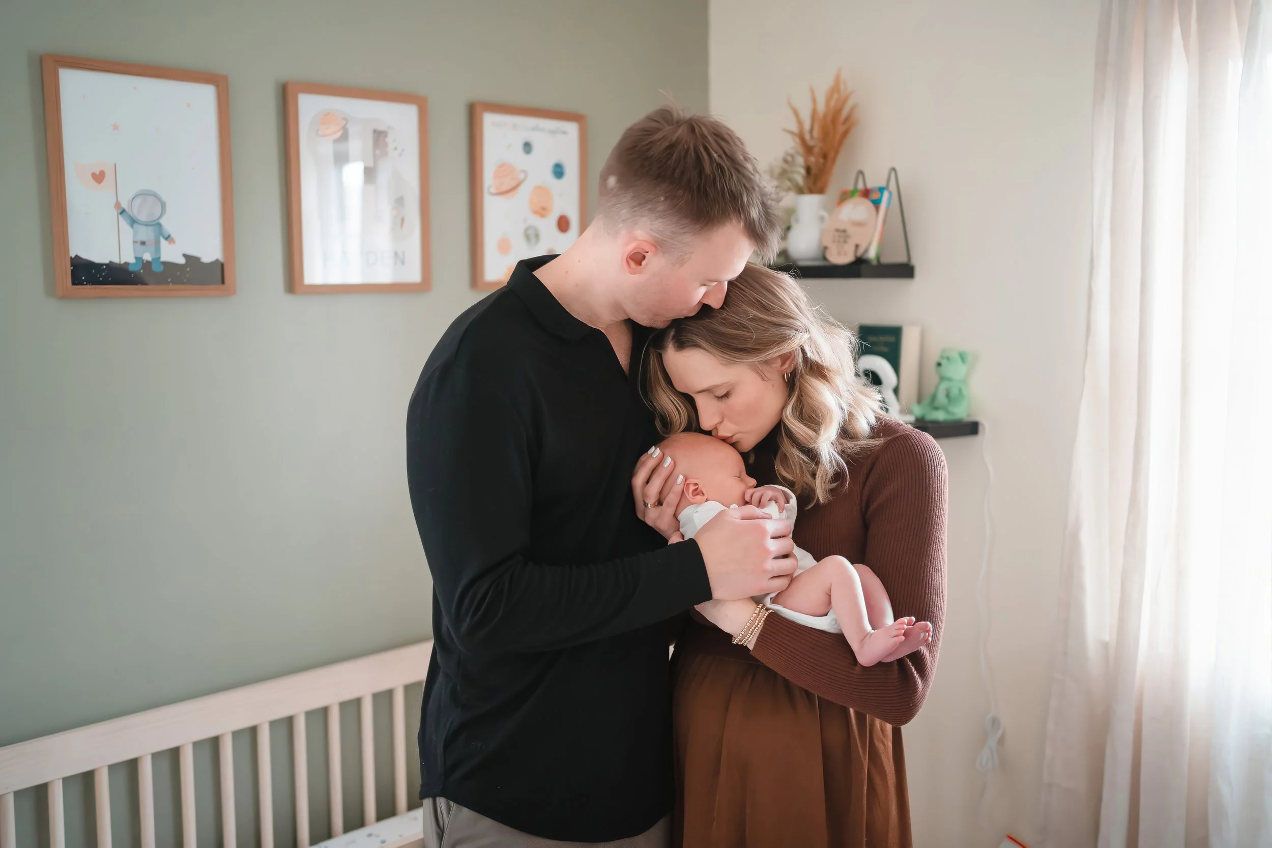 A man and woman holding and gazing at a newborn baby in a softly lit room with green walls and framed art, a shelf with decor, and a window with curtains.