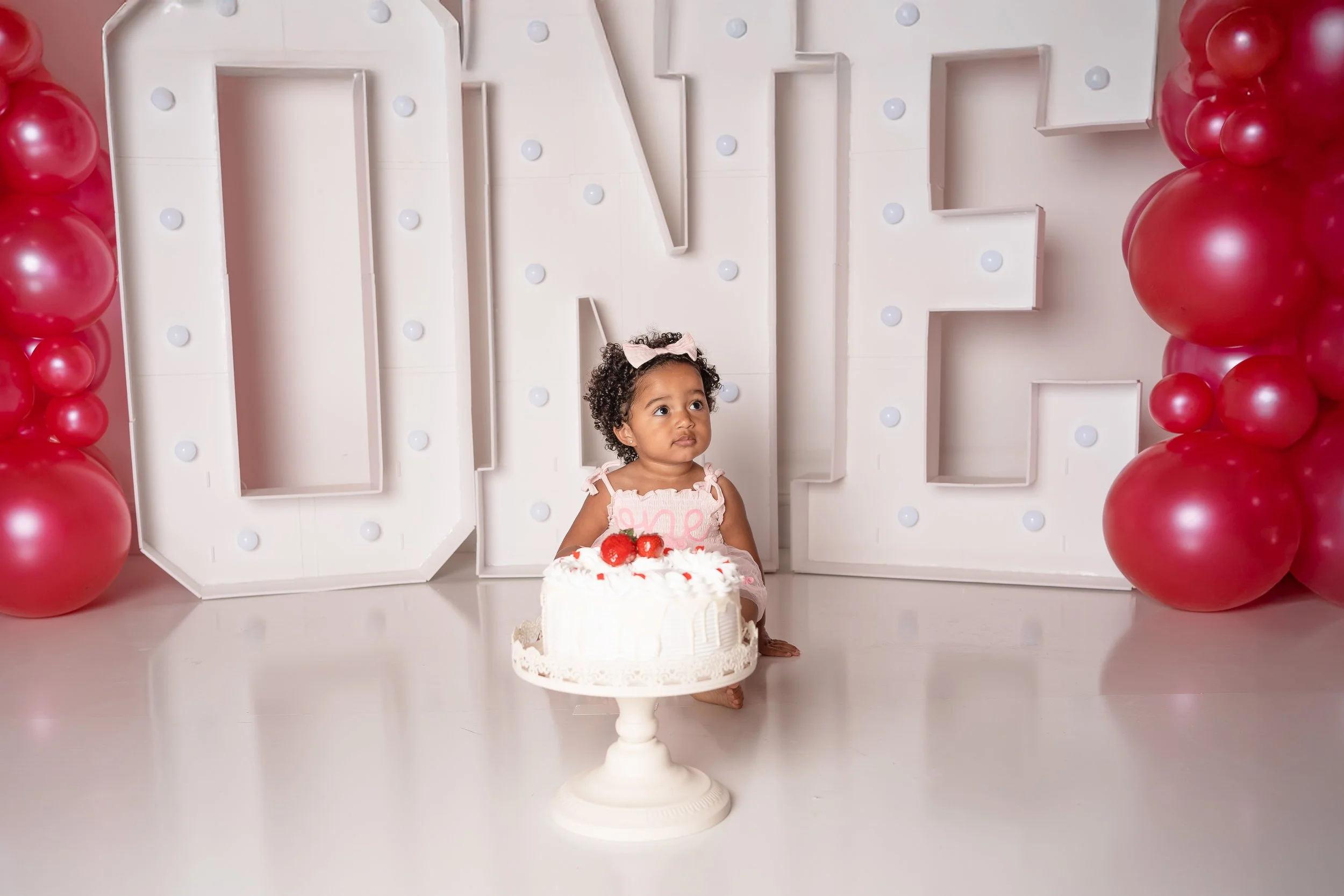 A young girl with curly hair and a pink bow in her hair, sitting on the floor, looks up, next to a white cake with strawberries on top, in front of a large decorative sign that spells out 'LOVE' surrounded by red balloons.