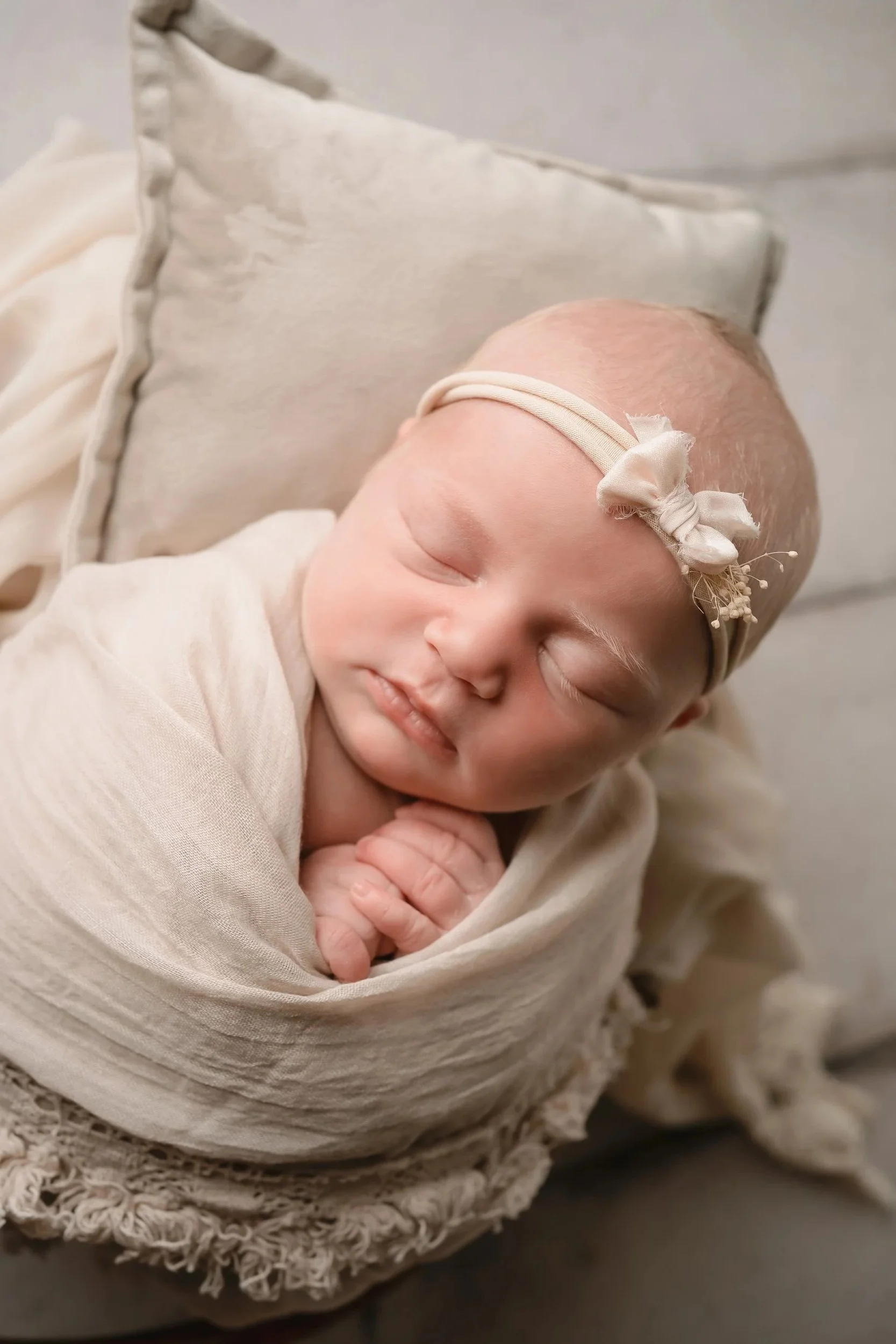 A sleeping baby wrapped in a soft beige blanket, wearing a cream-colored headband with a small bow, lying on a cream cushion.