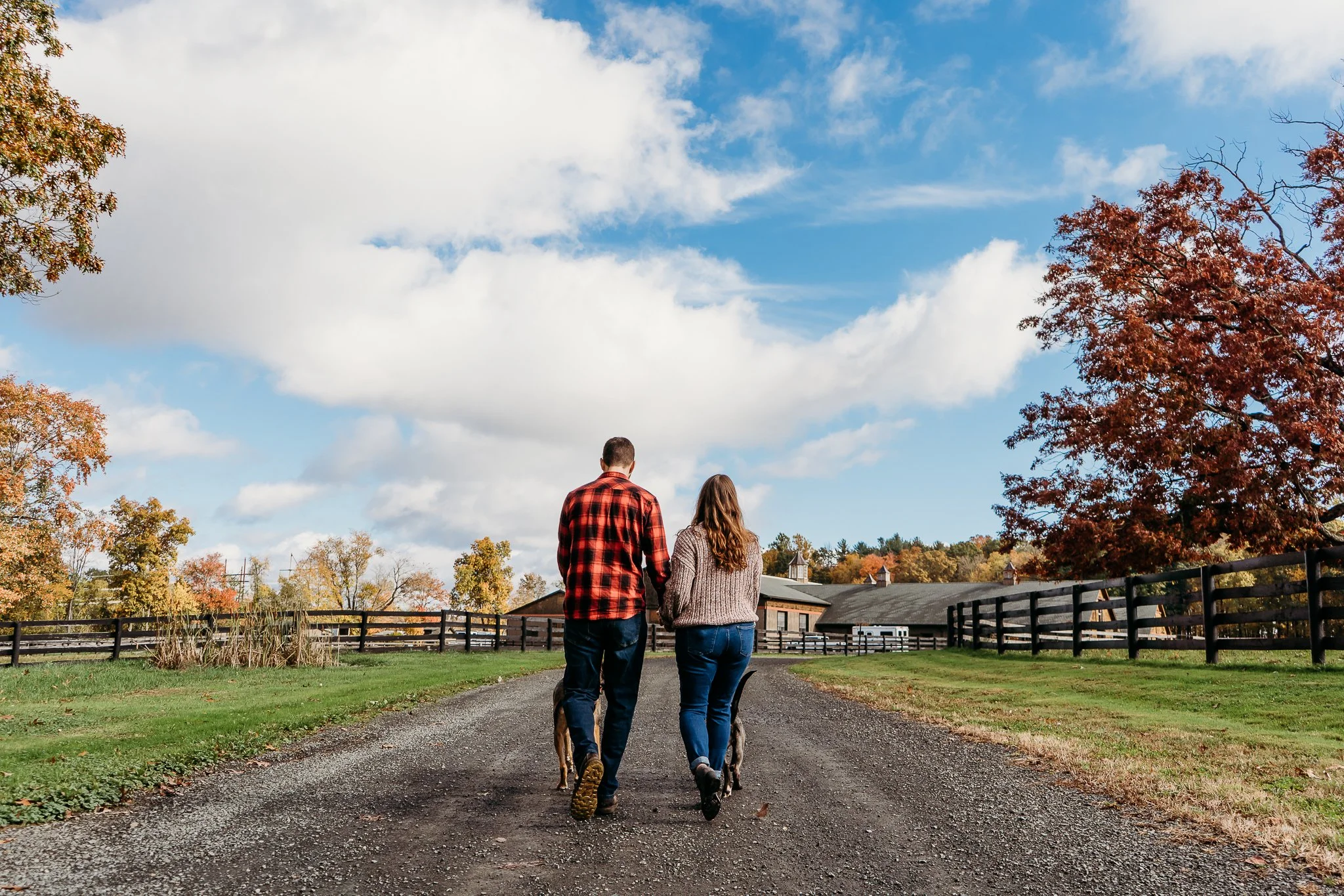 A man and woman walking with a dog in a park with trees displaying autumn colors, a fenced area, and a cloudy sky.