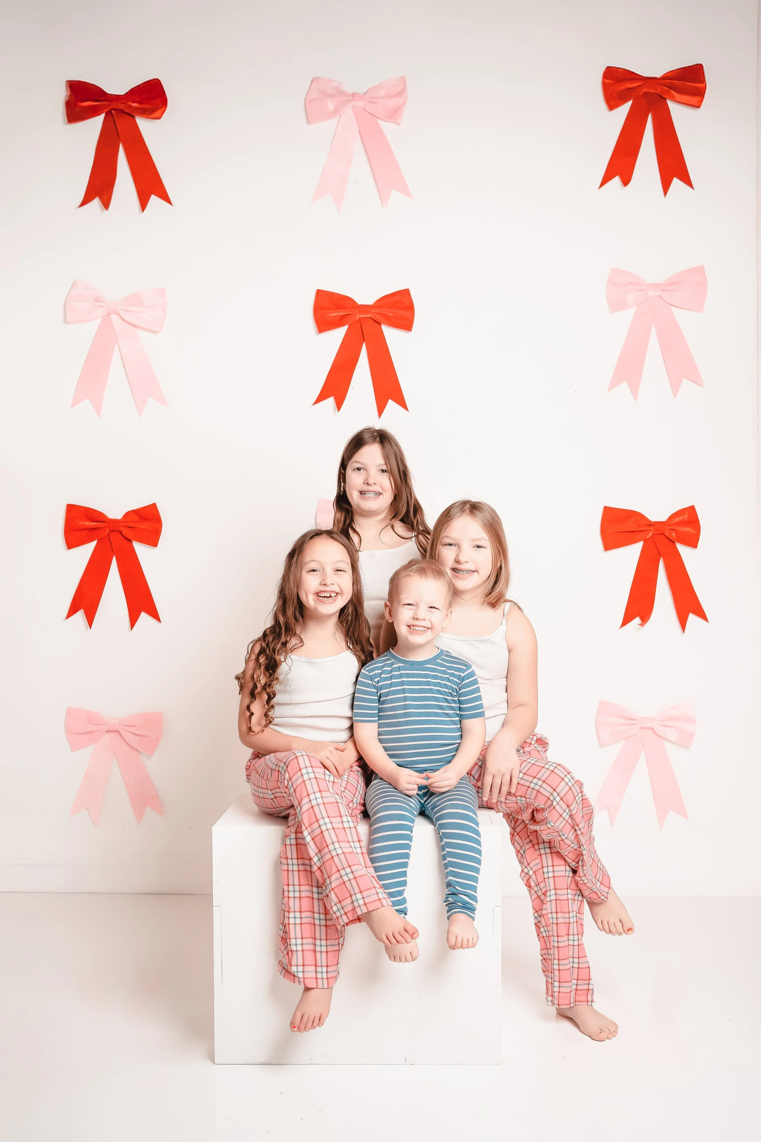 Four children sitting on a white box in pajamas, smiling, with a white background decorated with pink and red bows.